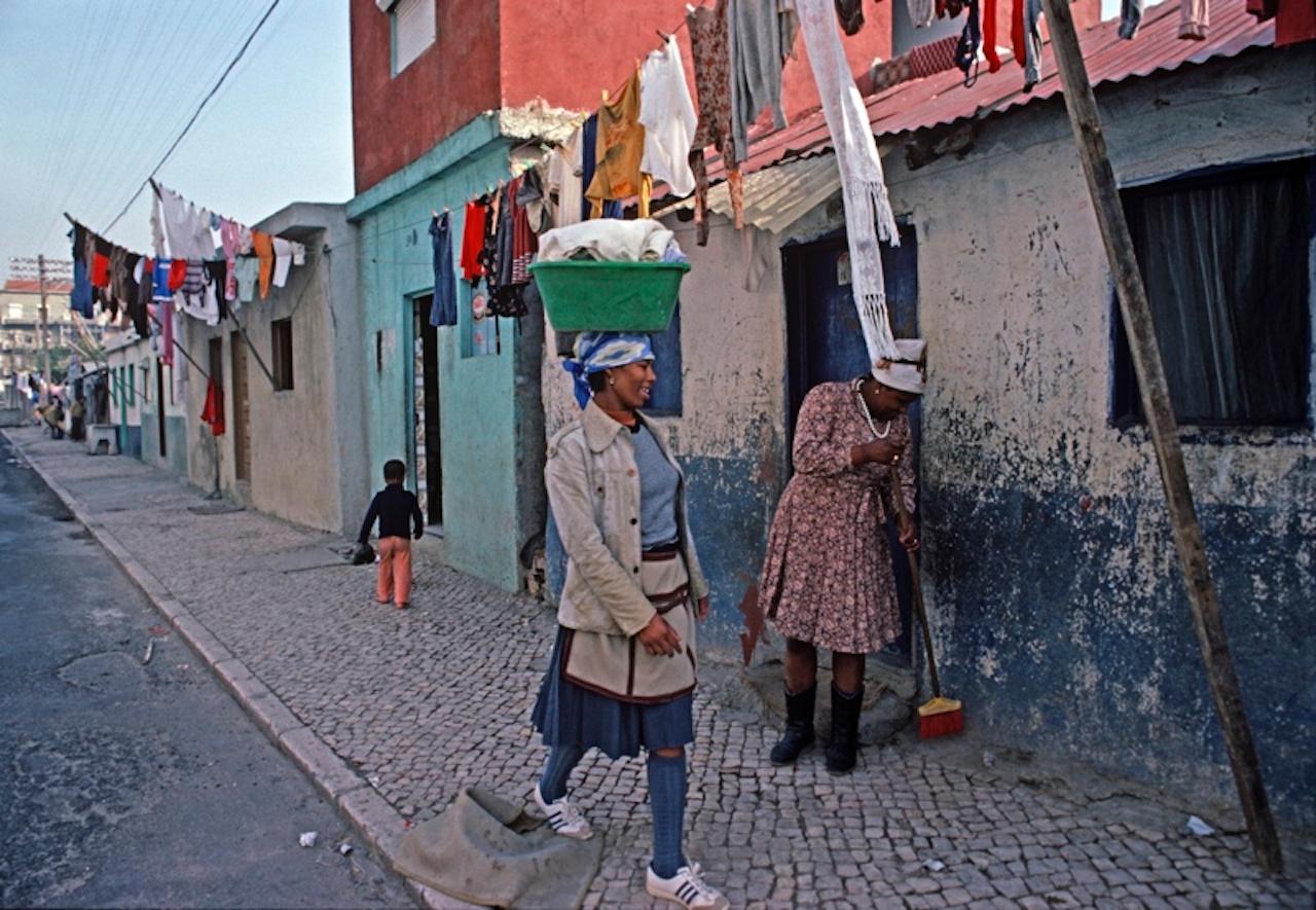Walk On By von Alain Le Garsmeur
Eine kapverdische Einwanderin, die einen Wäschekorb auf dem Kopf trägt, geht an einem Nachbarn vorbei, der den Bürgersteig vor ihrem Haus reinigt, Lissabon, Portugal, 1984.

Papierformat 16 x 20 Zoll / 40 x 50 cm