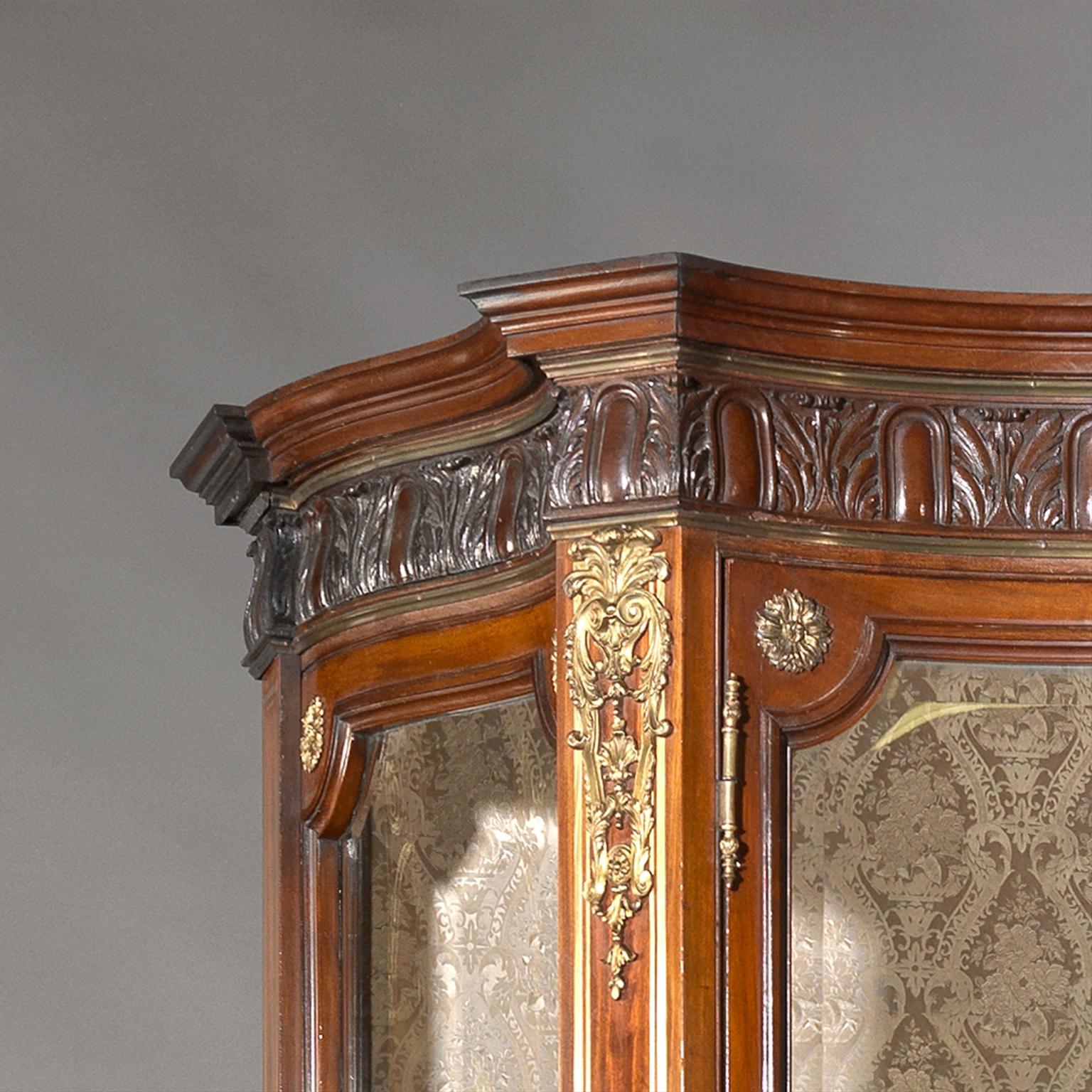 An impressive large gilt-bronze mounted mahogany vitrine.

This impressive vitrine has a shaped cresting rail carved with acanthus above a pair of shaped doors with bevelled glass panels and canted corners applied gilt-bronze flowerhead paterae.