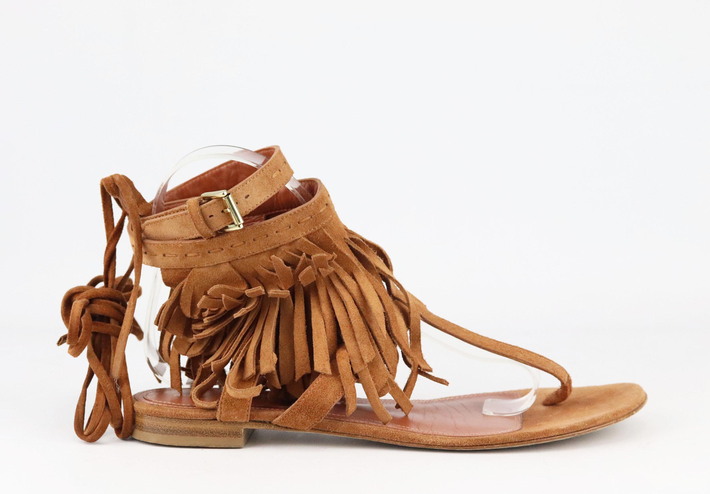 Las sandalias de Alberta Ferretti, confeccionadas en flexible ante color canela, están adornadas con los flecos característicos de la marca; este par tiene una favorecedora tira con doble hebilla en el tobillo y cordones inspirados en los