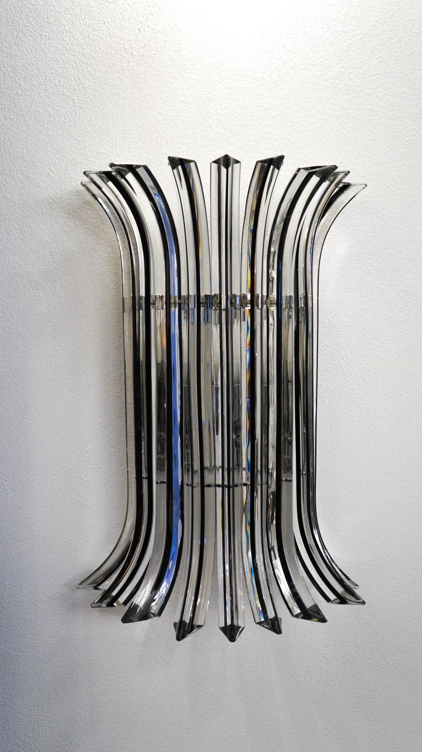 Eine sehr elegante Wandleuchte, die acht Glaselemente präsentiert, die in das Glas gebogen, aber mit
weiche und leichte Züge, verziert mit einer dünnen schwarzen Linie im Inneren des Glases, daher der Name des
farbe 