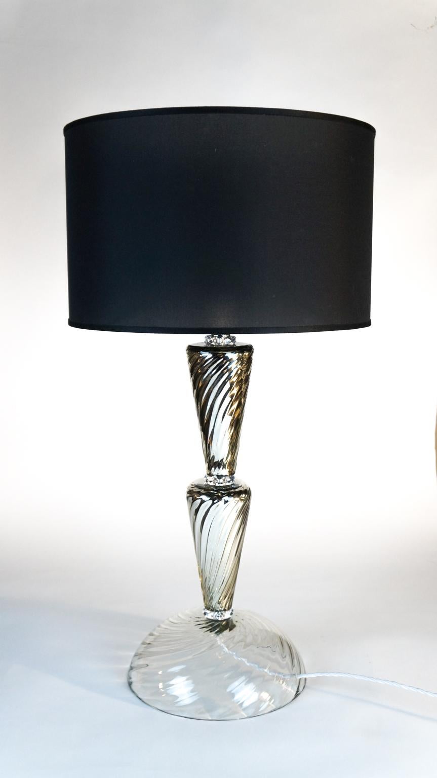 Soutenue par une importante base ronde de 30 cm de diamètre, cette lampe de table particulière se poursuit par deux cônes en miroir entre lesquels sont encadrés de petits cristaux qui créent un élégant jeu de lumière.
Projet du maître verrier de