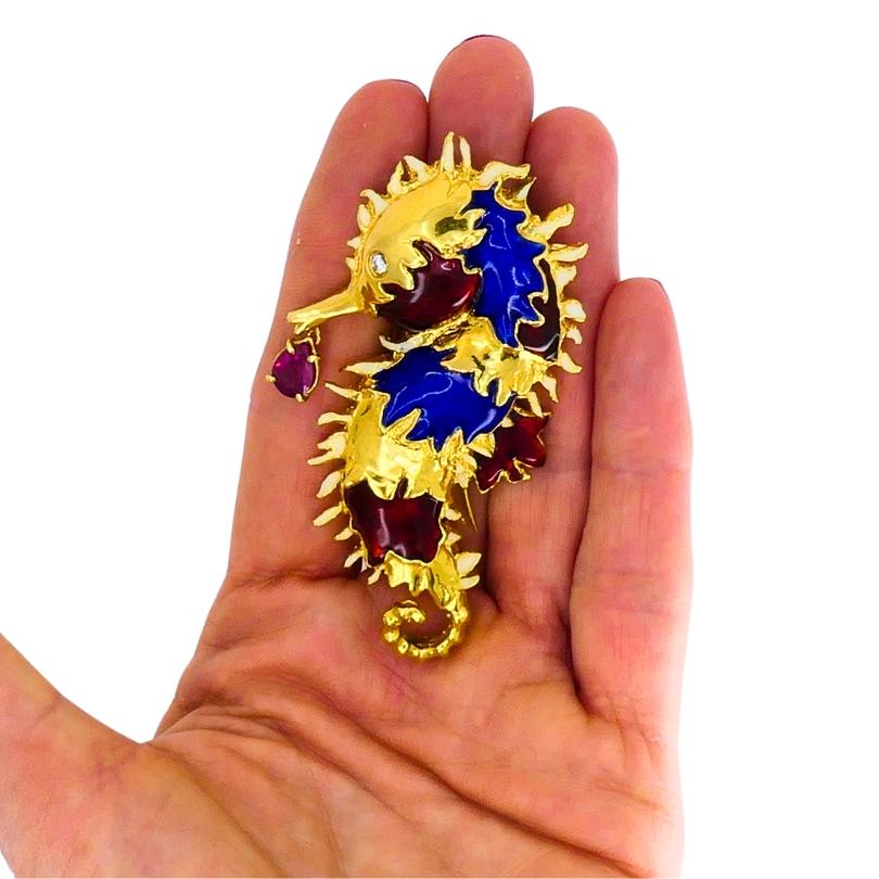 A vintage Aldo Cipullo for Cartier Seahorse brooch, made of 18k gold, featuring enamel and gemstones.
This charming Cipullo piece, with its textured gold body, features a playful mix of vibrant blue and deep red enamel. A tear-shaped ruby drop,
