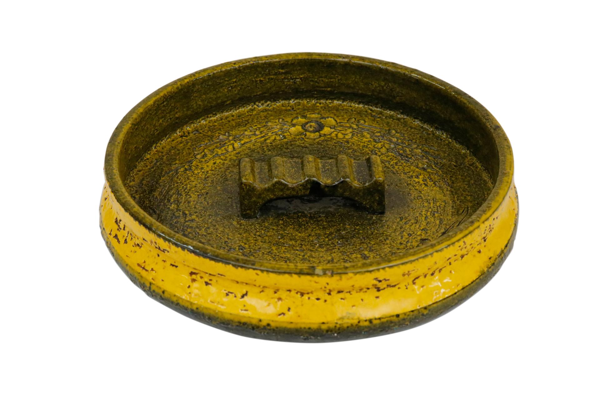 A 1950’s Aldo Londi for Bitossi Italian ceramic ashtray in yellow and olive green. Decorated with an incised floral spray motif. At the center is a raised cigarette holder. Numbered and signed “Italy” underneath.