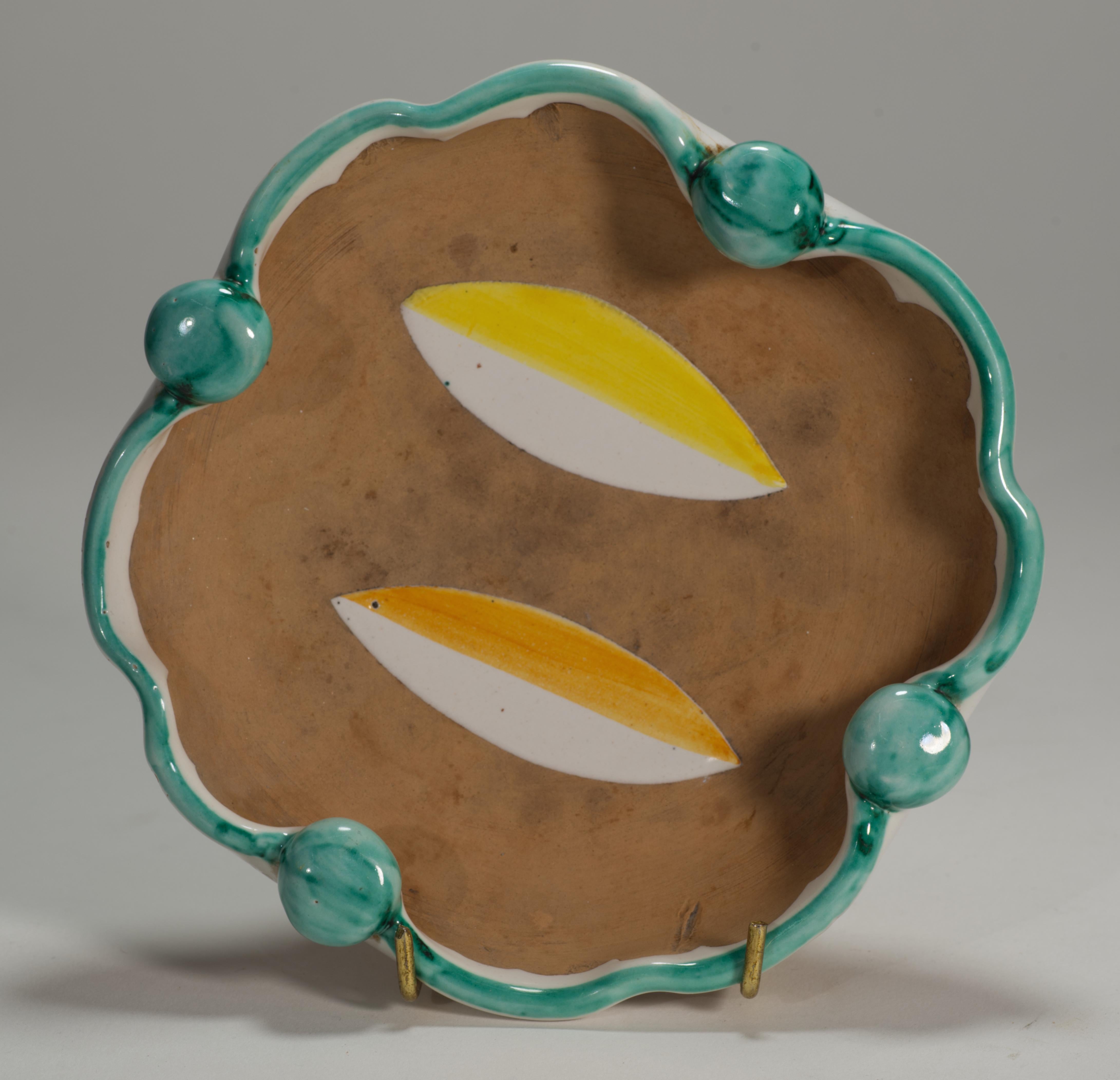  El cuenco o cenicero modernista de mayólica presenta una distintiva decoración de hojas blancas, amarillas y naranjas en el centro del cuenco, con el borde decorado en blanco y verde azulado. Una parte significativa del cuenco se deja sin esmaltar,
