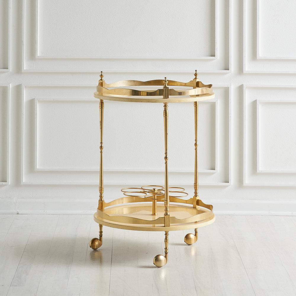 An 1950s Italian bar cart designed by Aldo Tura and produced in Milan. Featuring cream colored lacquered goatskin leather, gilded cast metal scalloped upper and lower railings, and finials and wine storage for 6 bottles. Includes original label to