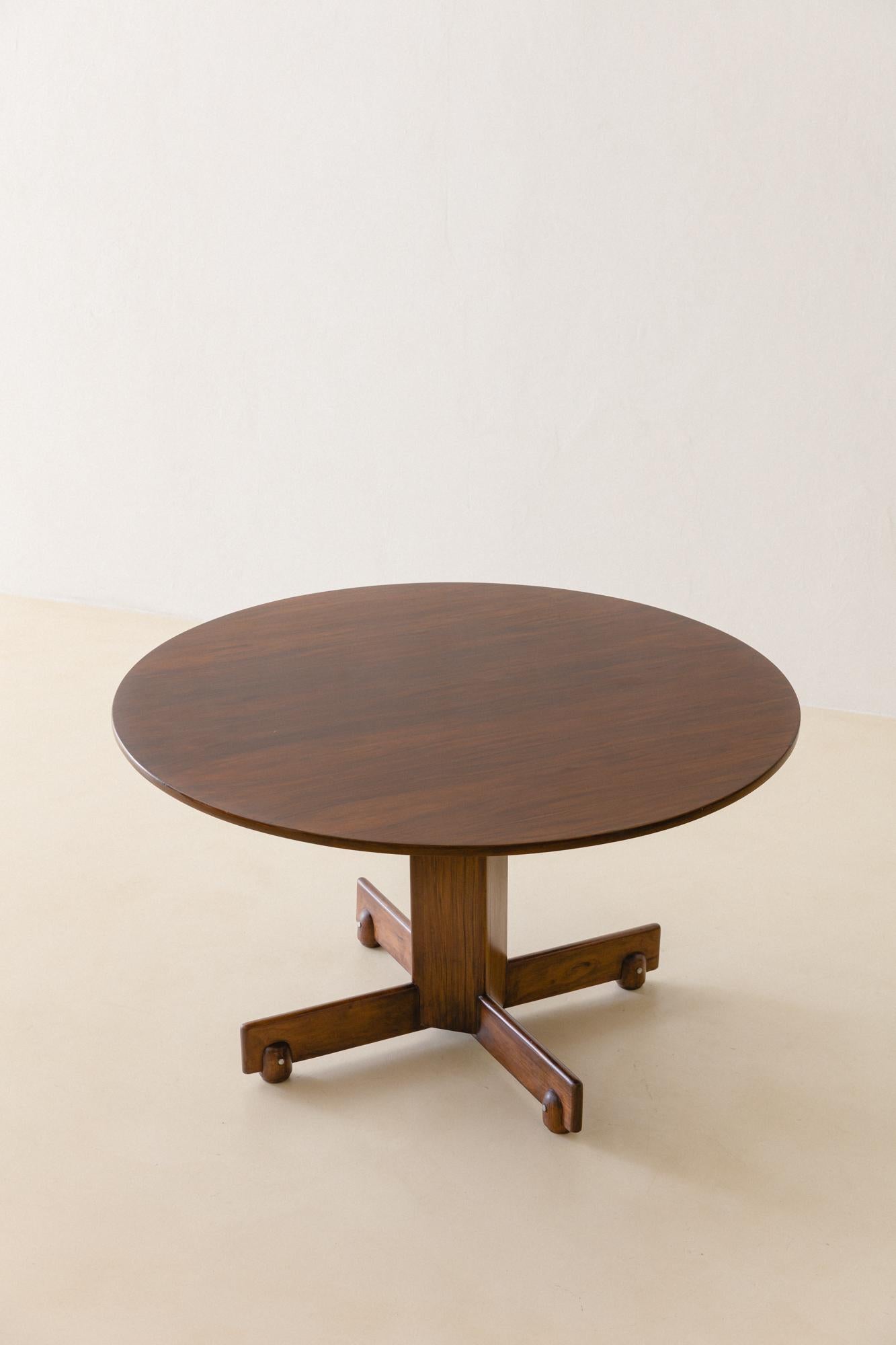 The Alex table is a rounded table made in solid Rosewood (Jacaranda), with both feet and top cross-shaped. 

Designed in 1960 by Sergio Rodrigues (1927-2014), this modern dining table features very well-built fittings, as the cross support for the