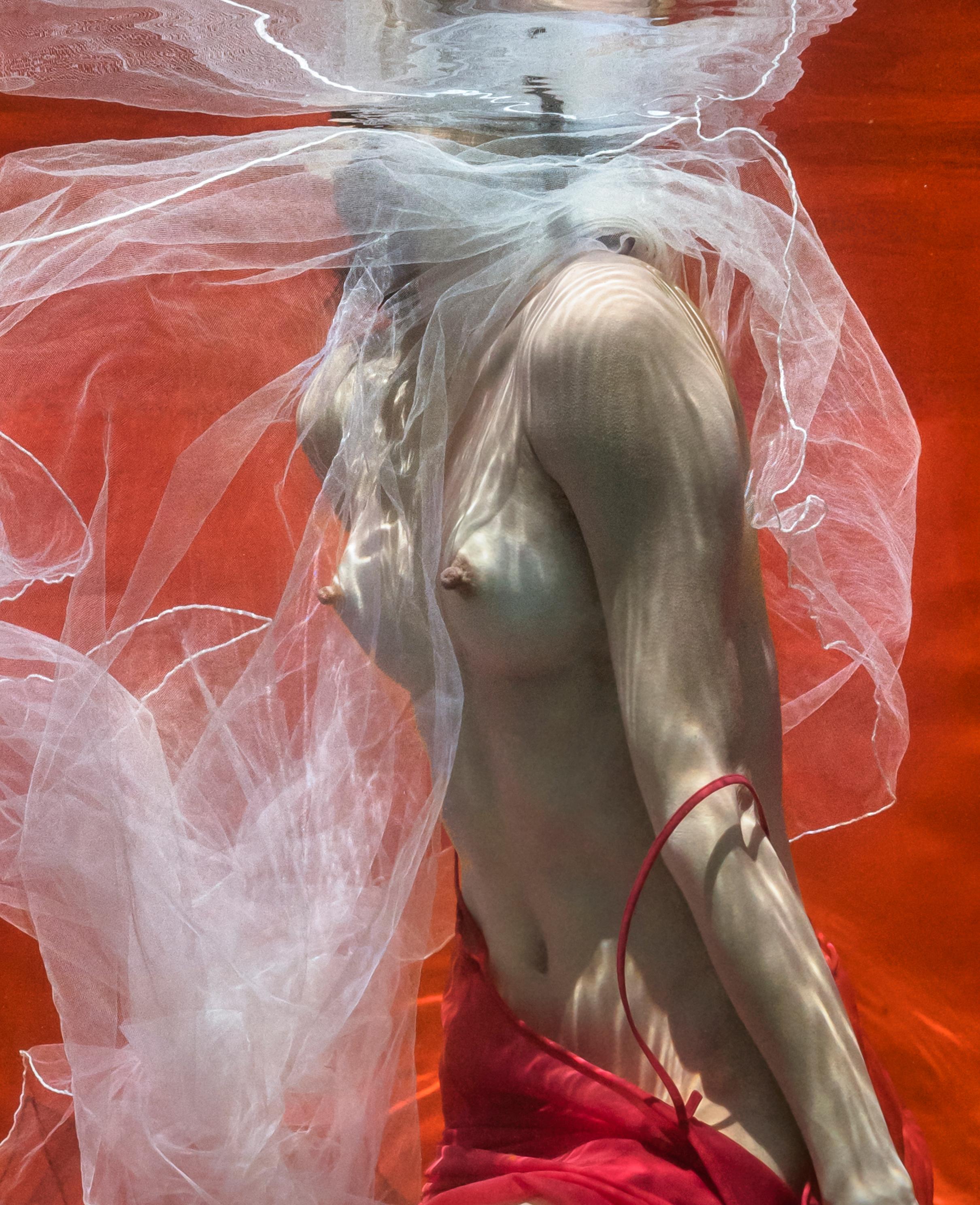 An underwater photograph of a topless young woman with white tulle scarf taking off her red skirt on bright red background. 

Original digital print on archival paper signed by the artist.
Limited edition of 24 print 2
Paper size: 62x44