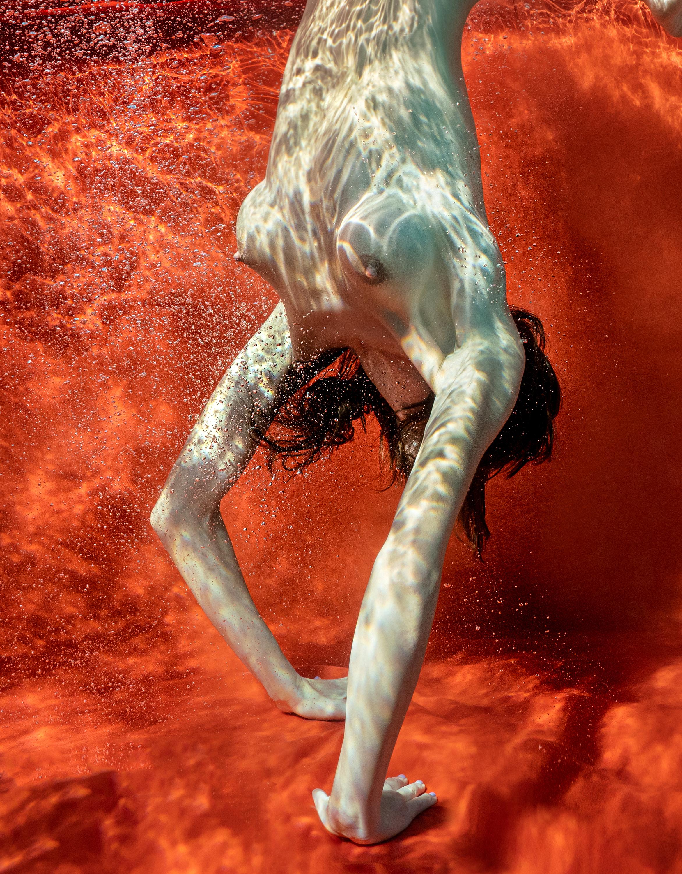 Underwater photograph of a naked young woman standing on her hands on the bottom of a pool. Her perfect body on the red background and classic heavy breasts leave spectators speechless. It is particularly true for the big prints 44 by 58