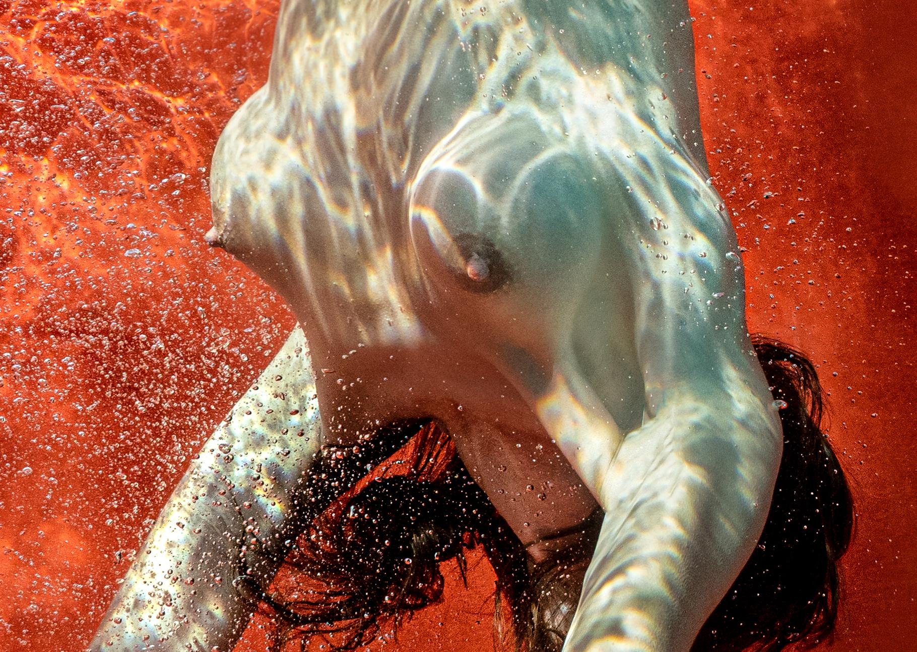 Unterwasseraufnahme einer nackten jungen Frau, die auf ihren Händen auf dem Grund eines Schwimmbeckens steht. Ihr perfekter Körper auf dem roten Hintergrund und ihre klassisch schweren Brüste machen die Zuschauer sprachlos. Das gilt insbesondere für