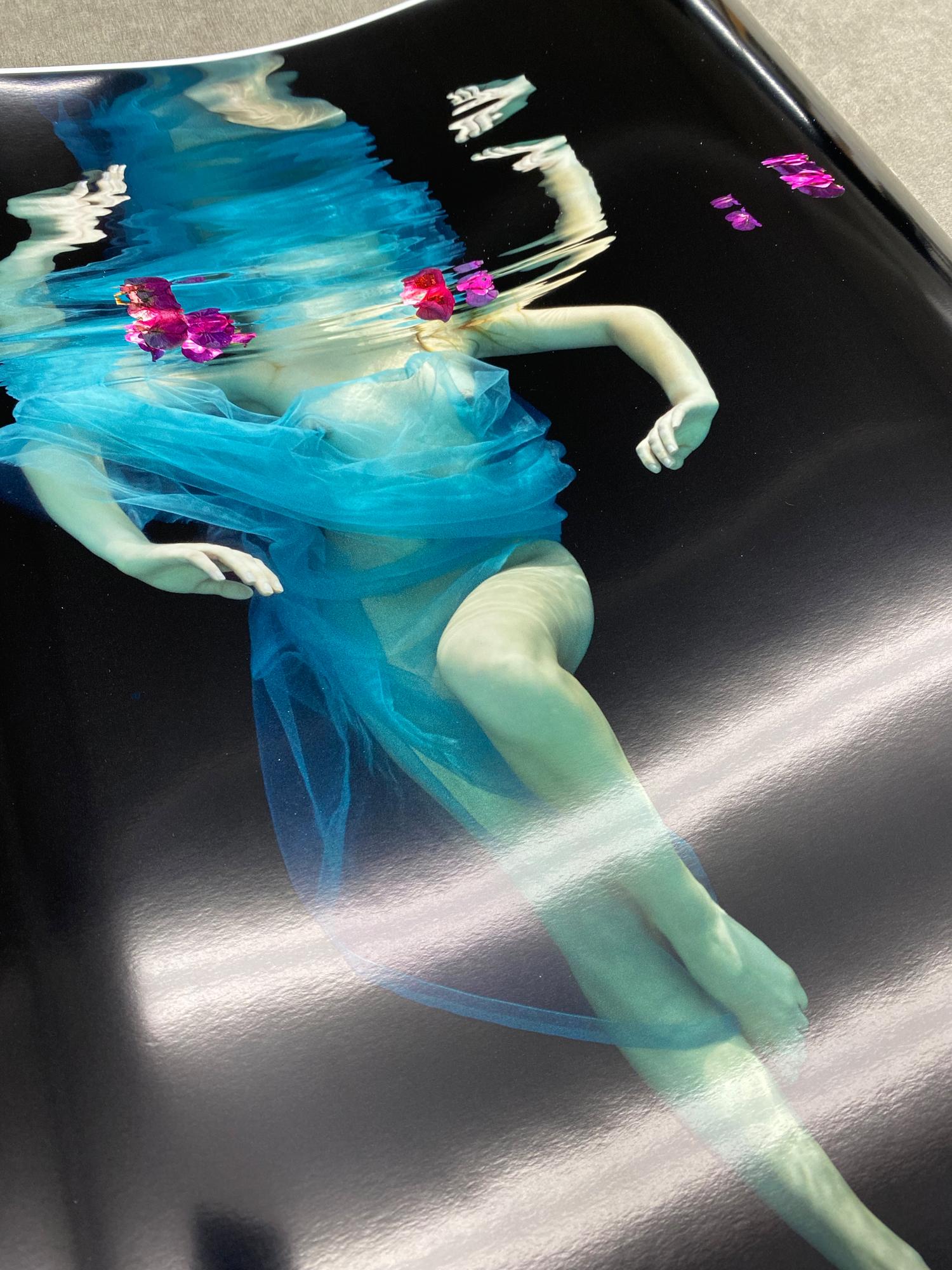 This ethereal underwater photograph captures a topless female figure draped in flowing turquoise tulle, suspended against a dramatic black backdrop. Vibrant magenta flowers float at the water's surface, creating striking color contrasts with the