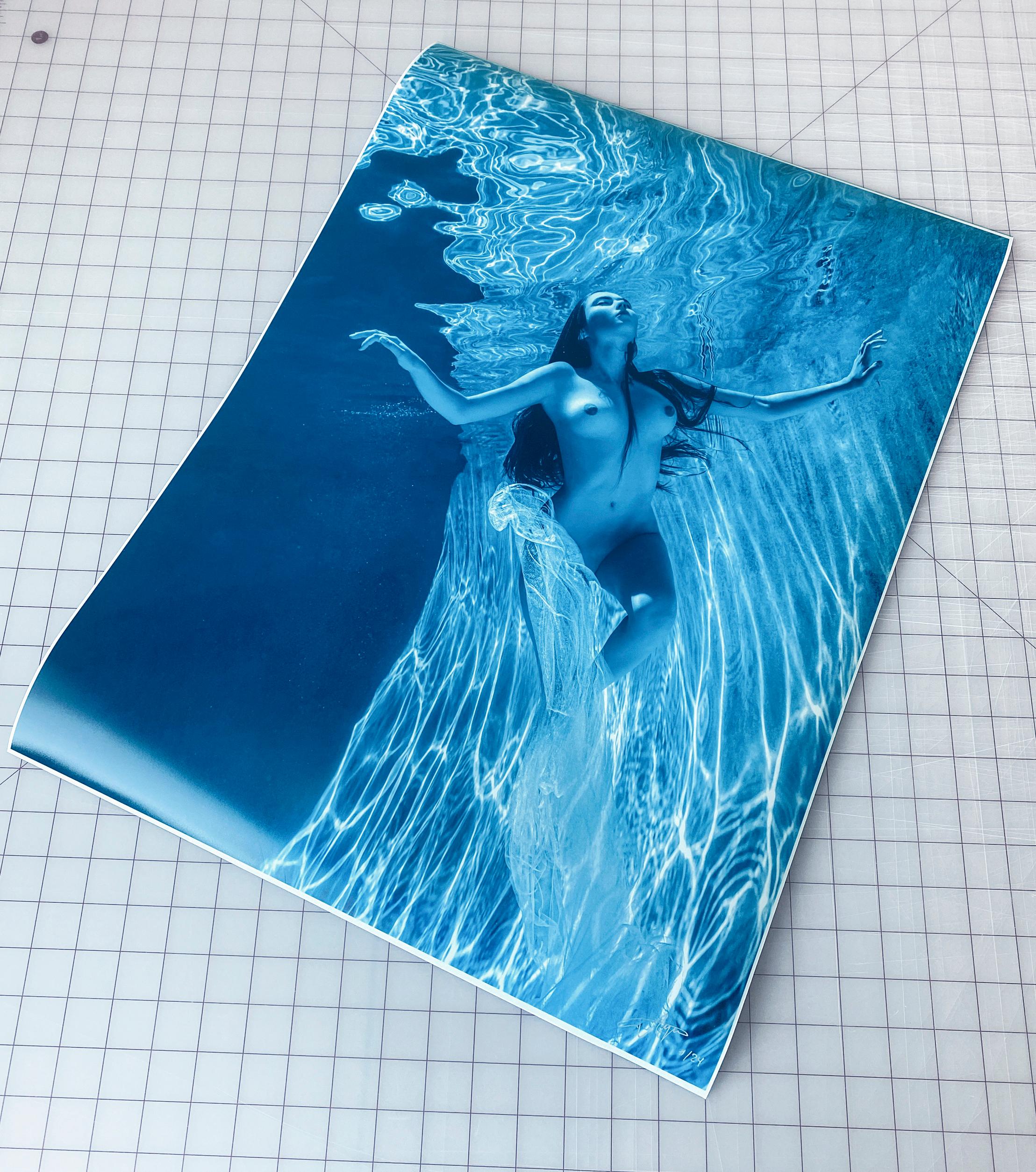 Photographie sous-marine d'une jeune femme nue aux cheveux bruns dans une piscine.  Une écharpe en soie transparente glissait sur ses épaules, ouvrant les seins et couvrant partiellement les hanches et le pubis. Il s'agit d'une photographie