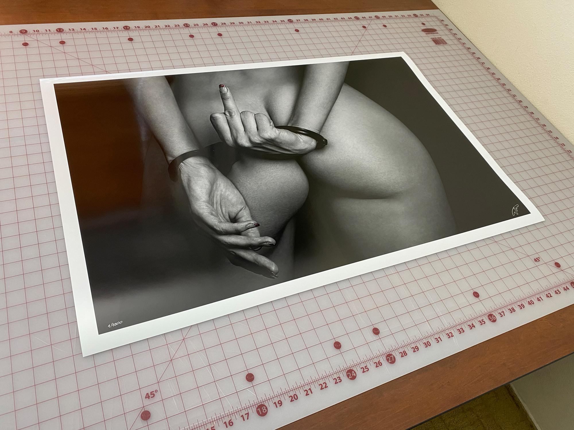 A black and white photograph of a perfect woman's buttocks and her handcuffed hands giving a finger.

Original gallery quality archival pigment print signed by the artist. 
Limited edition of 1000. Print #3.
Paper size: 23x36