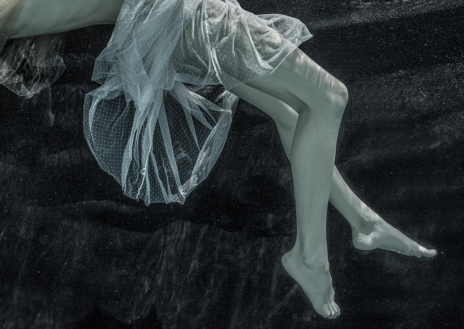 Unterwasser-Schwarz-Weiß-Fotografie einer jungen Frau, die anmutig unter der dunklen Wasseroberfläche balanciert. Ihr Tüllkleid und ihre Spiegelung auf der Unterseite des Wassers bilden eine exquisite Komposition, die an die elegante Wölbung eines