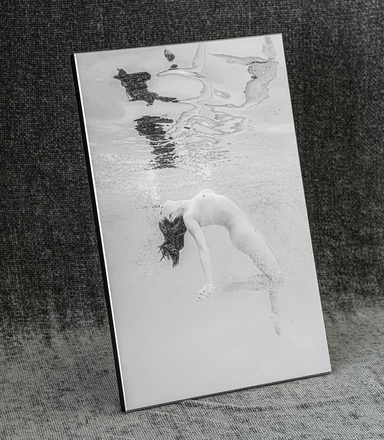A black and white nude photograph of a young woman diving in a pool. The woman is coming from the depth up toward the surface and her body reflects in the internal water surface.

Original print on aluminum plate with solid black backboard. The