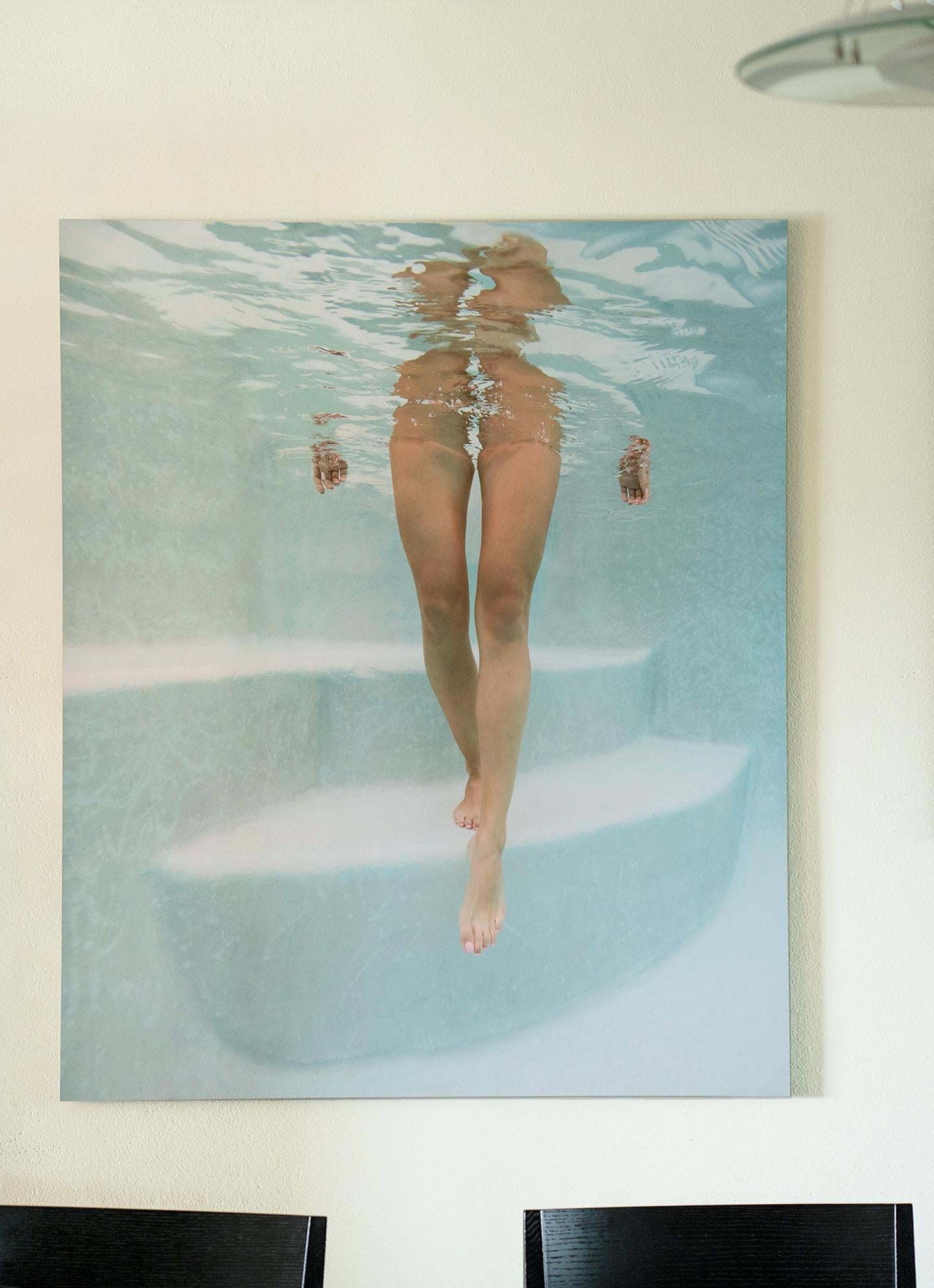 Une photographie sous-marine légère et très seyante d'une fille descendant les marches de la piscine vers vous. La photo bleu clair ne montre aucun maillot de bain ni aucun endroit où l'on pourrait s'attendre à un maillot de bain. Cette photographie
