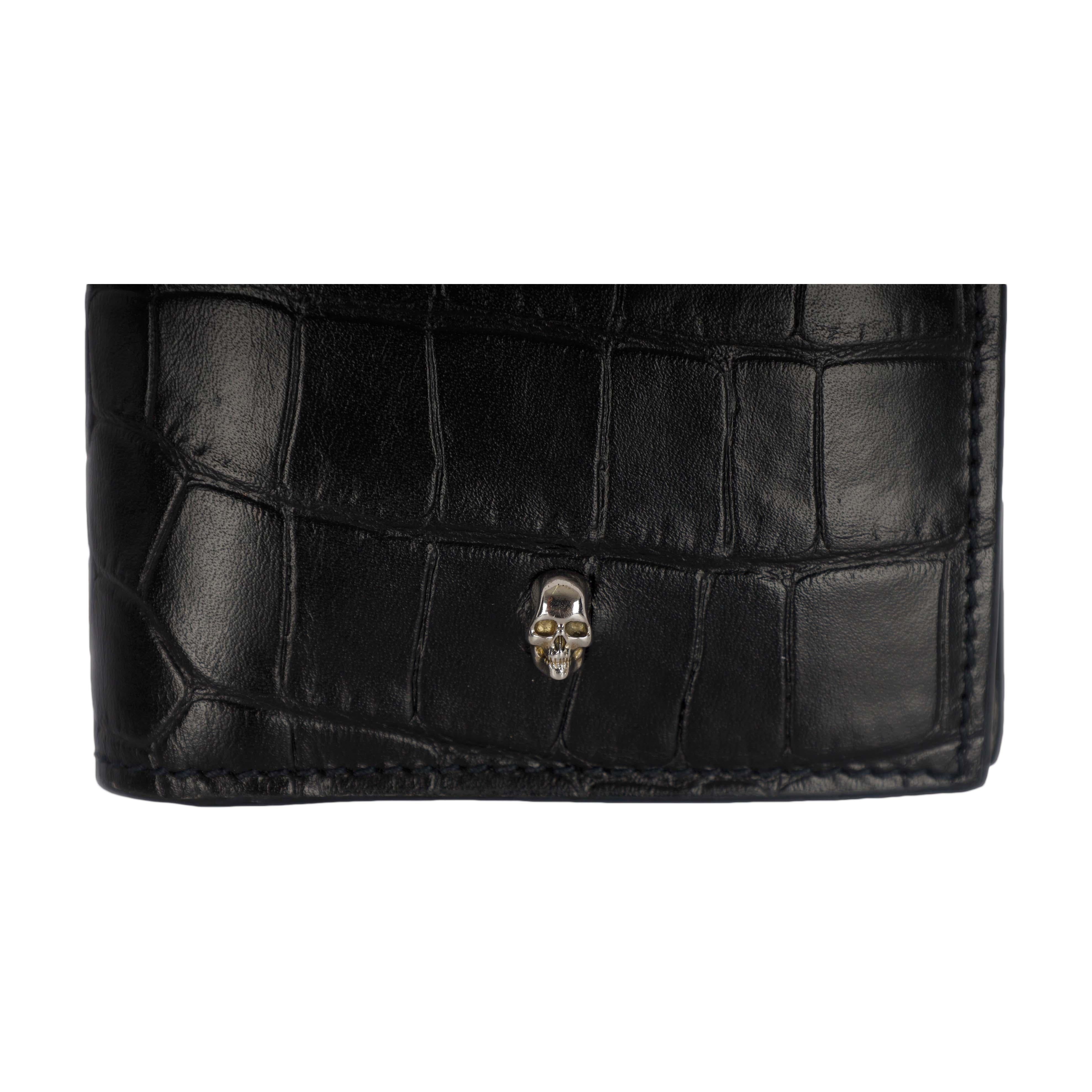 The Alexander McQueen black rectangular folded wallet with silver-hued skull features on embossed crocodile leather effect on the outside and blue card slots with a zip-up coin pouch and note compartment on the inside. Made in Italy, this piece in