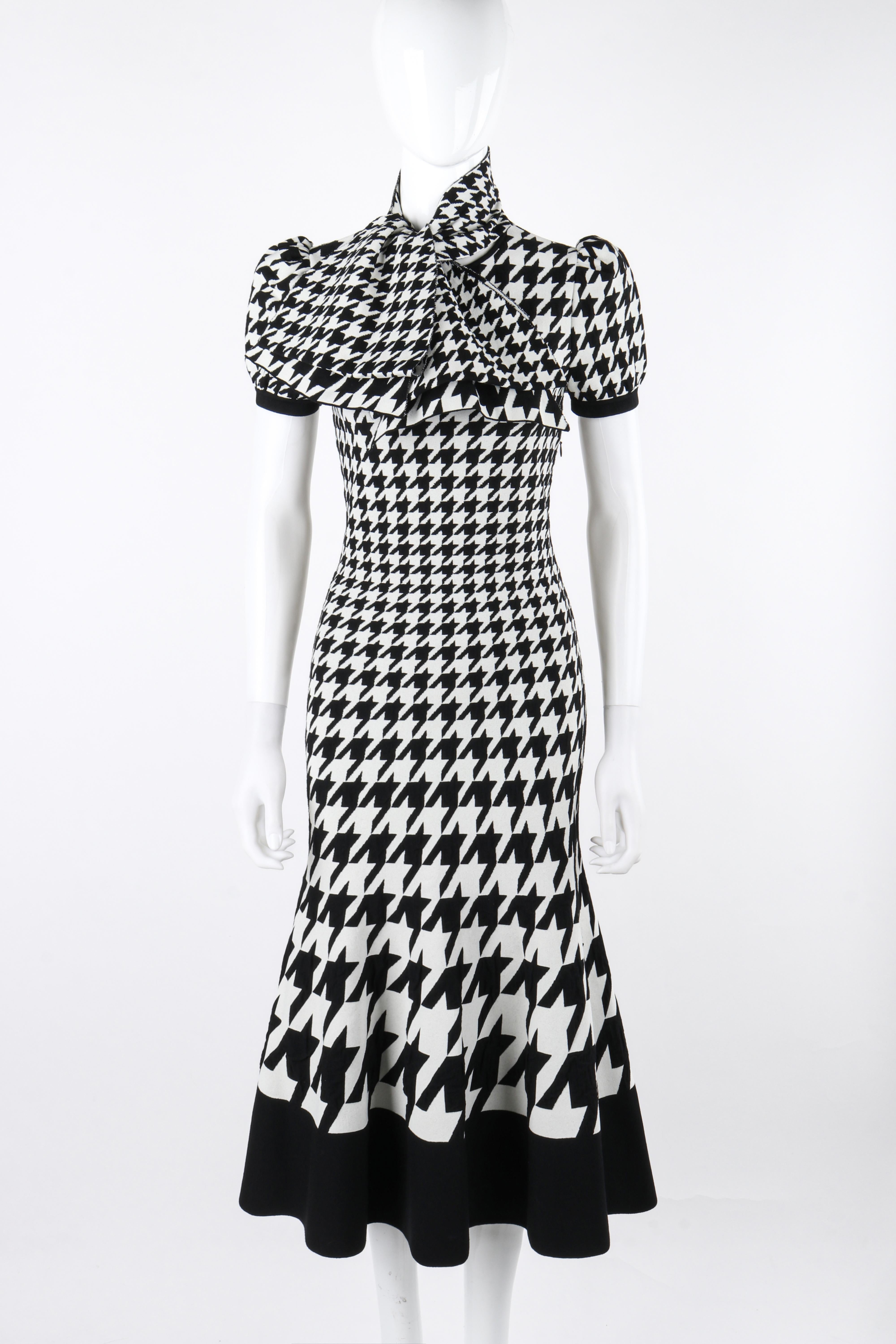 This houndstooth calf length dress by Alexander McQueen for the fall/winter 2019 collection features a form fitting design with a round neckline, short puffed sleeves, a full skirt with contrasting black trim, and an oversized coordinating neck tie