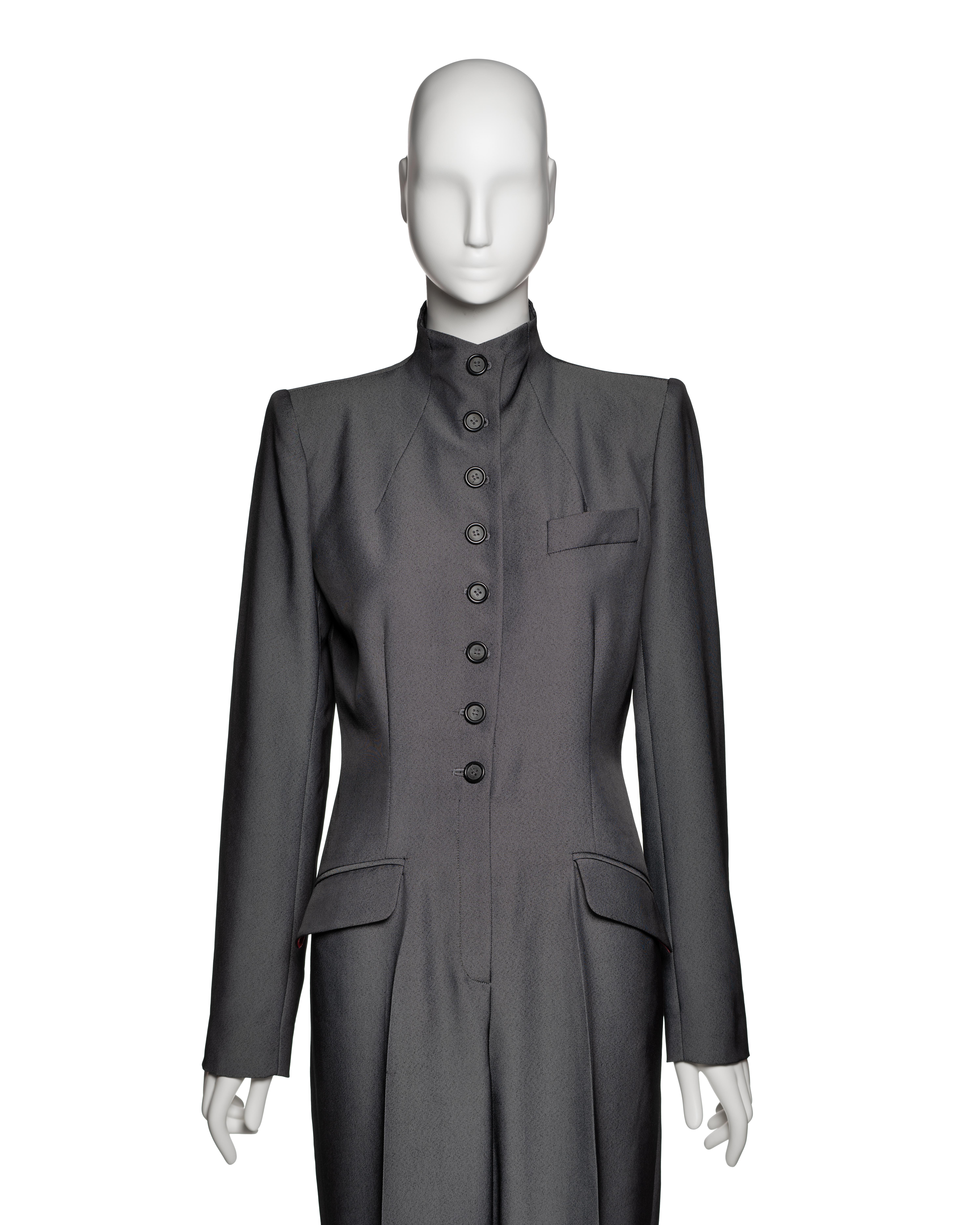 Tuta sartoriale monopezzo di Alexander McQueen della collezione Autunno-Inverno 1998, realizzata in twill di lana grigio con una leggera lucentezza. Il design è direttamente influenzato dalle uniformi militari storiche, in particolare dai cappotti