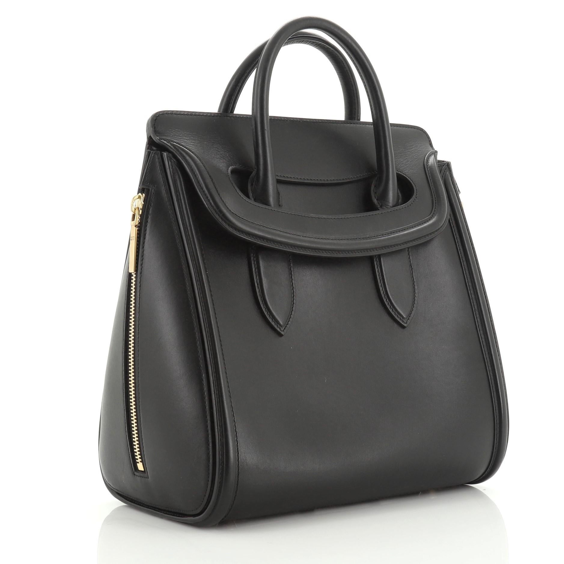 This Alexander McQueen Heroine Tote Leather Large, crafted from black leather, features dual rolled leather handles, gusseted side zipper details, protective base studs, and aged gold-tone hardware. Its flap closure opens to a gray suede interior