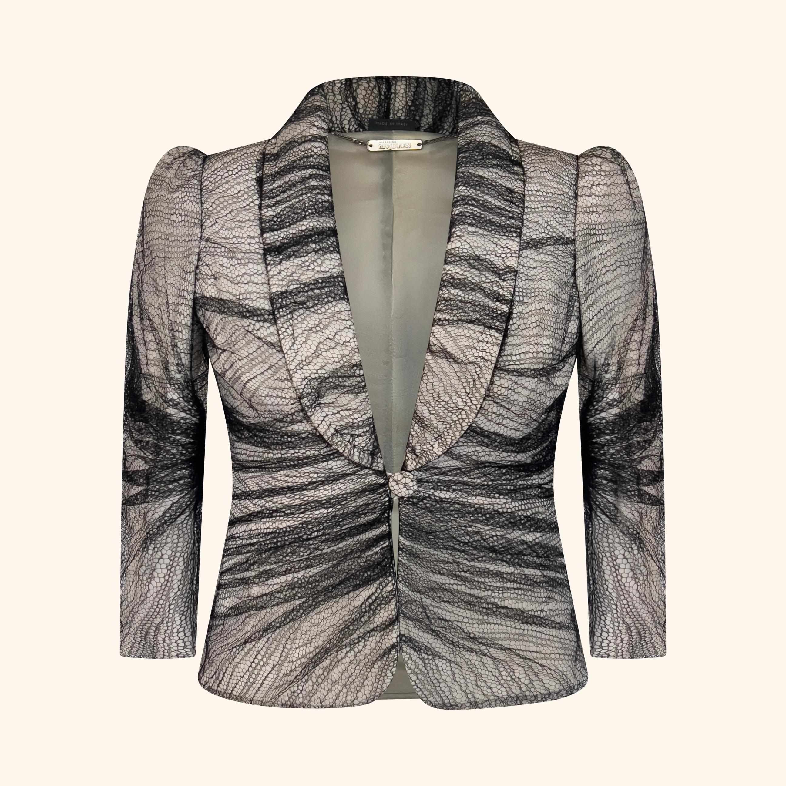 Il delicato pizzo nero è stratificato, plissettato e drappeggiato su una base di seta bianca, creando un contrasto strutturato.
La giacca è tagliata a monopetto, incorniciata da un bavero a scialle e da spalle a pagoda accentuate.
Rifinito con una