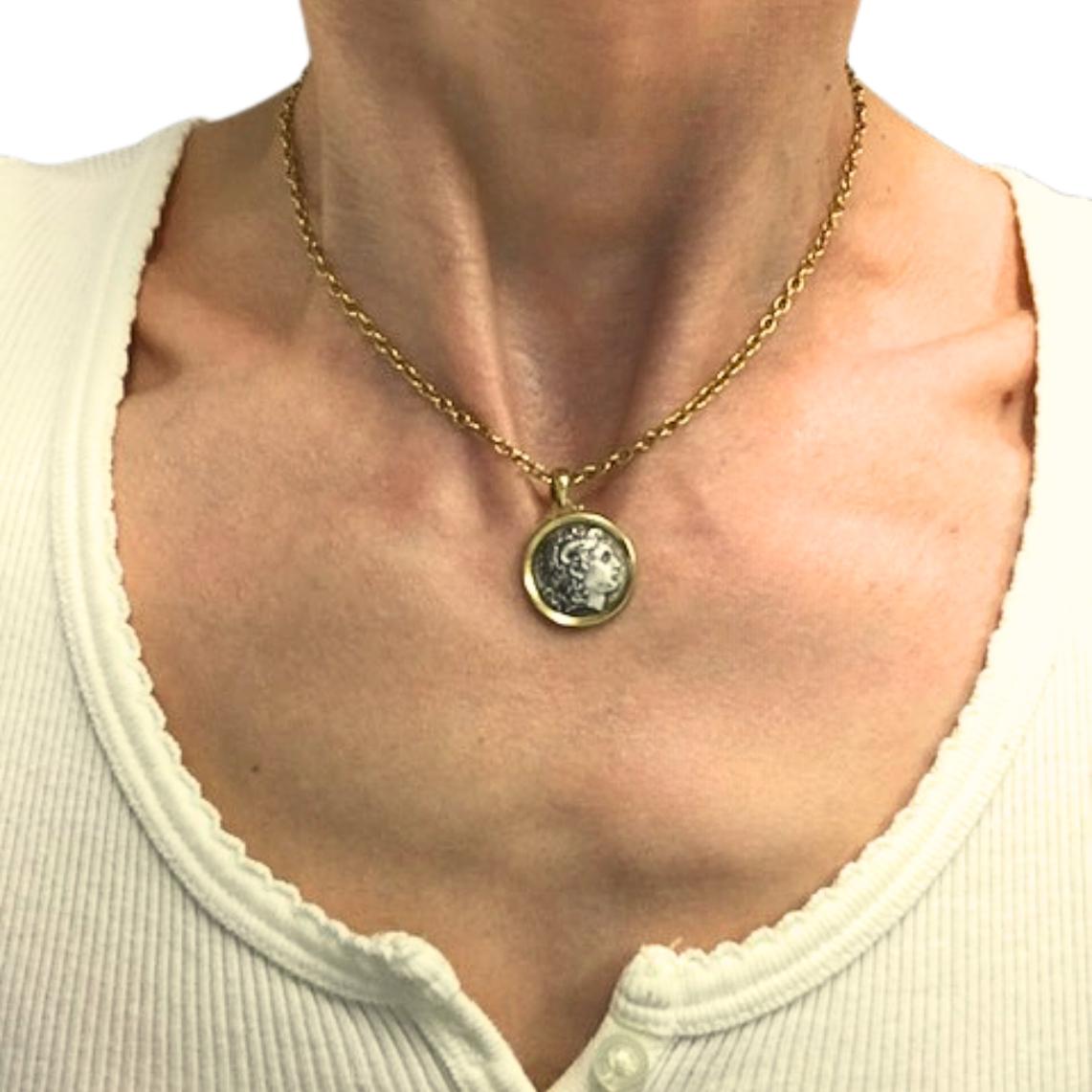 A vintage necklace, featuring a silver Alexander The Great coin charm set in 18k gold and a 14k cold chain necklace.
The obverse side of the coin, shown in the full necklace image, depicts Alexander the Great. The image is framed by a smooth gold