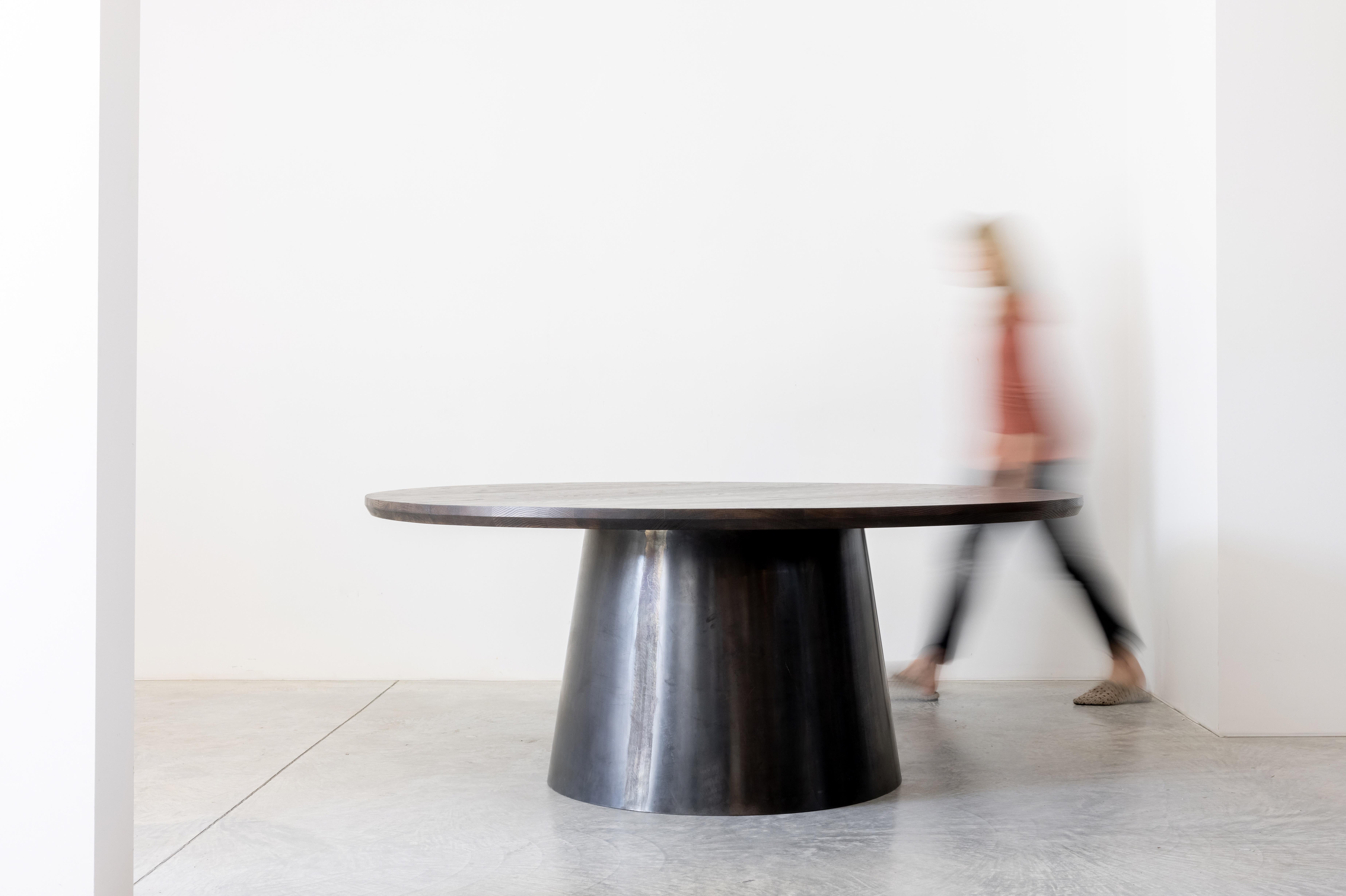 Ein dramatischer runder Tisch mit einem dicken Fuß aus geschwärztem Stahl. Der gebogene, konische Sockel ist sorgfältig gekrümmt und so zusammengefügt, dass eine fast nahtlose Verbindung entsteht, die als skulpturale Form wirkt. Die Verjüngung um 8