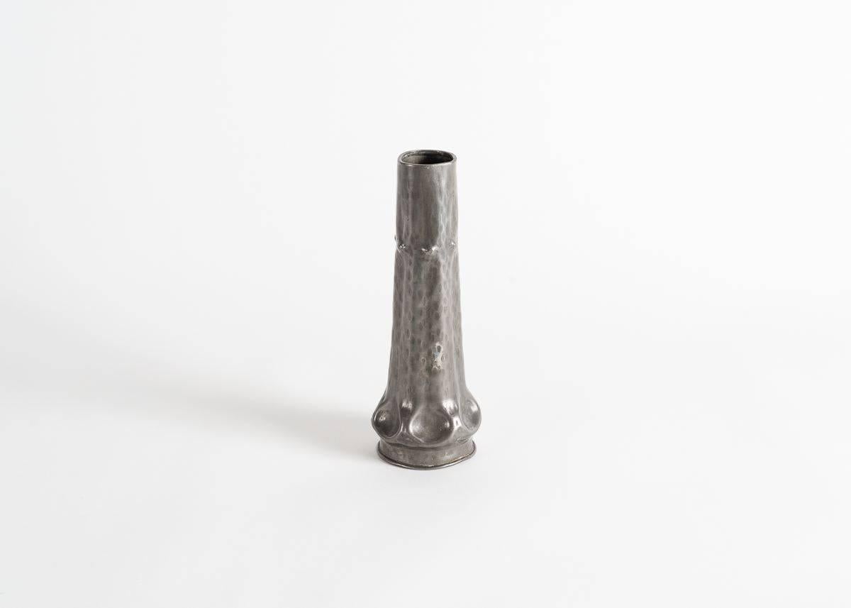 Firmato: A.E. Chanal

Un piccolo vaso tubolare in metallo, sottilmente accentuato da rientranze arrotondate sulla base e da protuberanze appuntite intorno alla bocca.
 