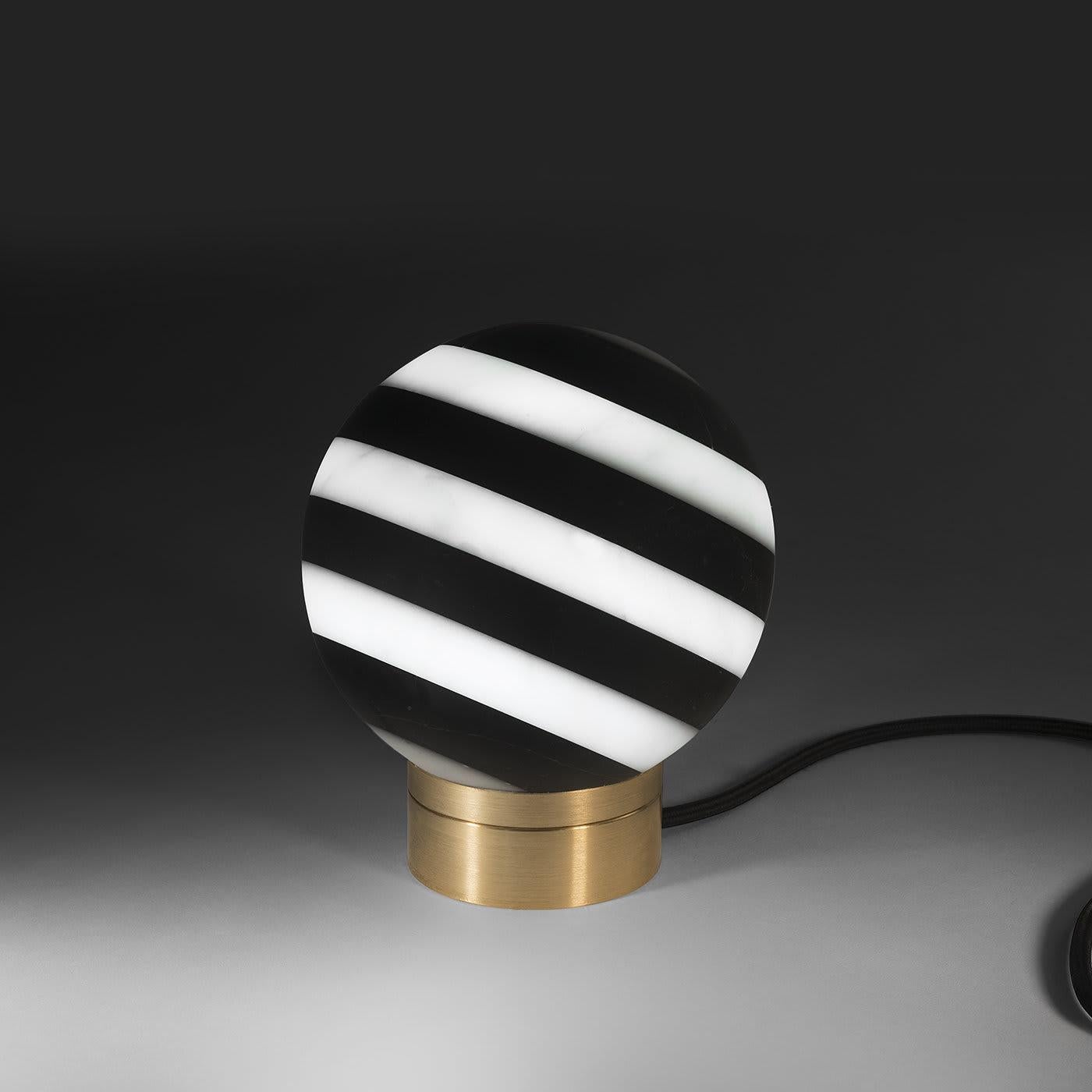 Inspired by the captivating black and white stone buildings found across Europe, this exceptional table lamp will cast an alluring glow in a traditional or minimalist decor. Mounted on a circular brass base with a gold finish, the orb-shaped lamp is