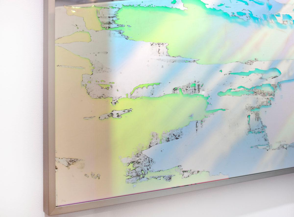 This large abstract fine art mirror by Alina B is made with dichroic film on mirror, and is framed in a champagne-toned frame. The film on the mirror allows for it, and the space it reflects, to appear to change colors to pink, orange, green, and