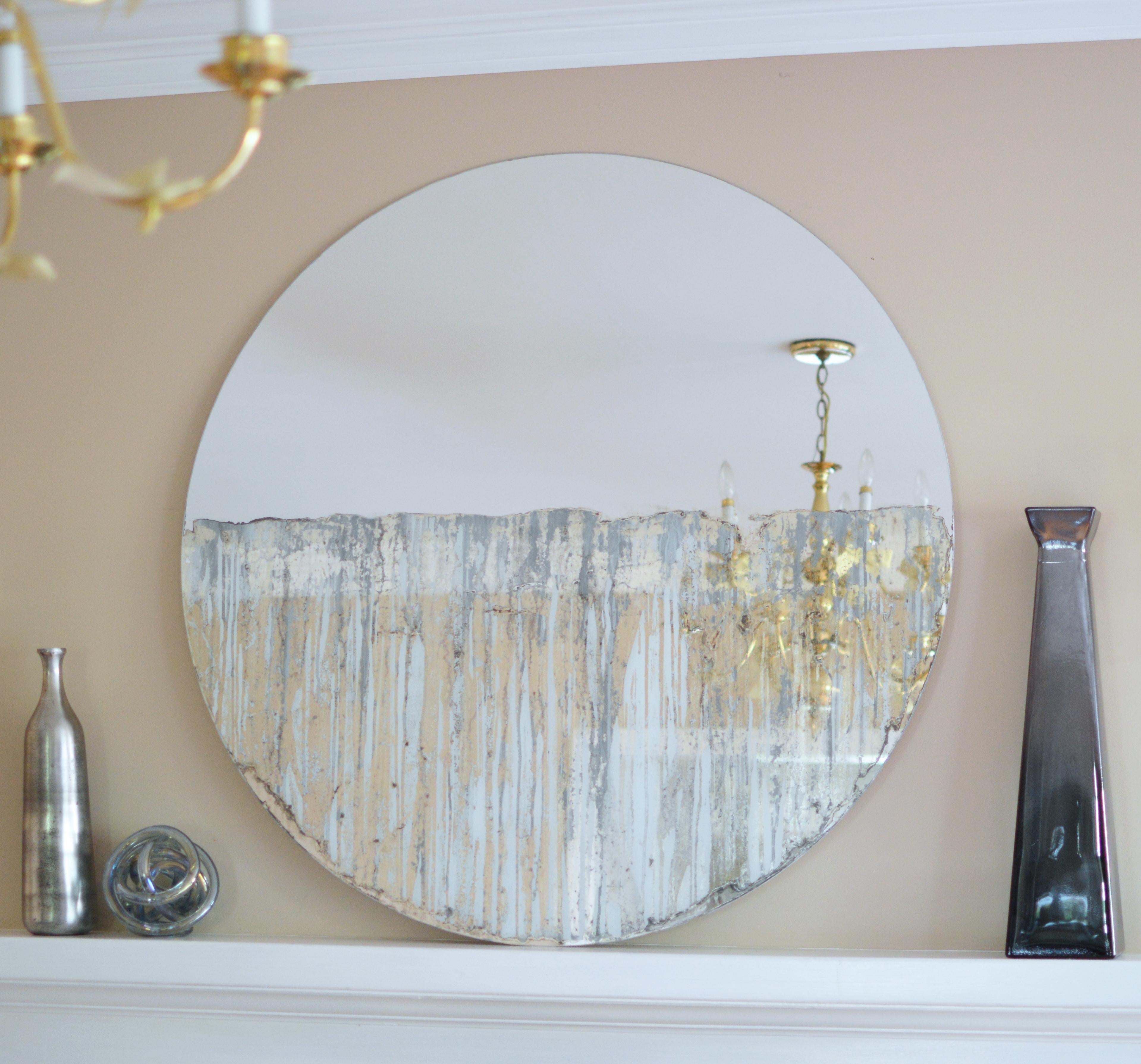 This contemporary mixed media piece is made with mixed media on a circular mirror. Deep blue-grey color has been applied over the bottom half of the mirror, giving it a painterly, dripped aesthetic but also allowing for mirror to continue to show