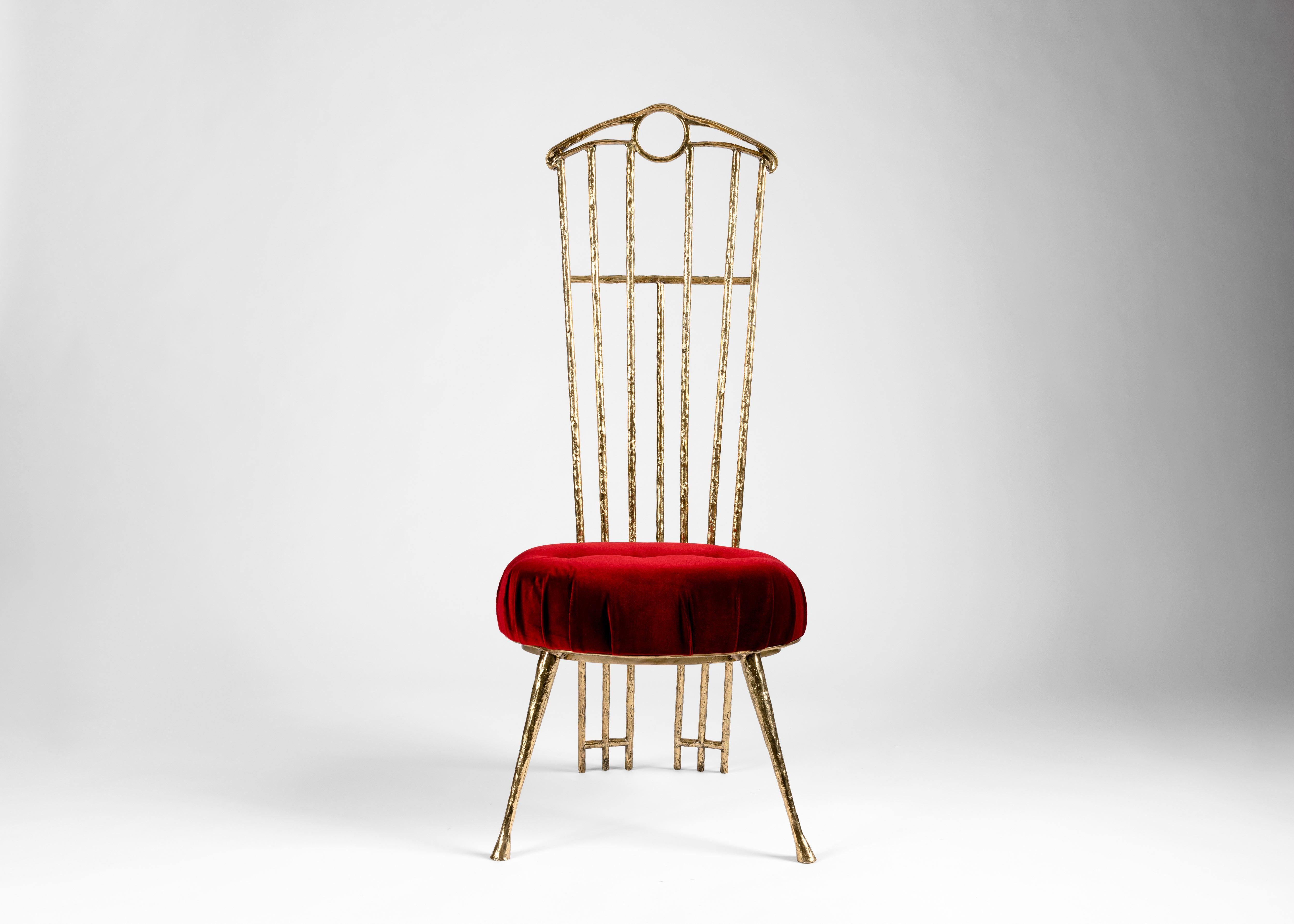 This striking piece is a playful take on formality. The chair's polished bronze, its rigid back, and its red upholstered cushion all suggest a throne, yet it rests upon playful, splayed legs, is executed in compositional lines of wavering
