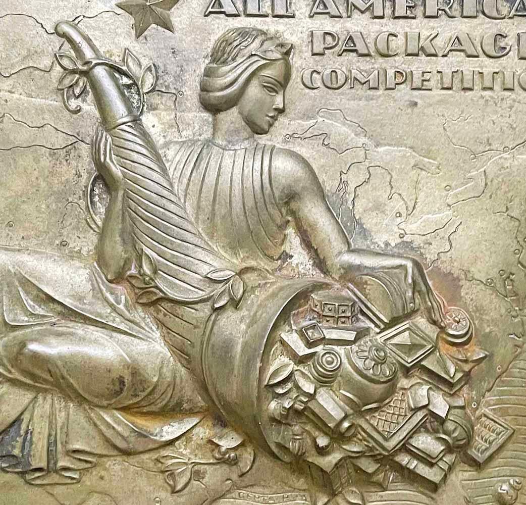 Gorgeously cast in bronze, given a pale gold patina, and set in a walnut plaque, this quintessential Art Deco bas relief presents a delicious conceit: Its classical figure is holding a cornucopia overflowing not with the produce of the earth, but