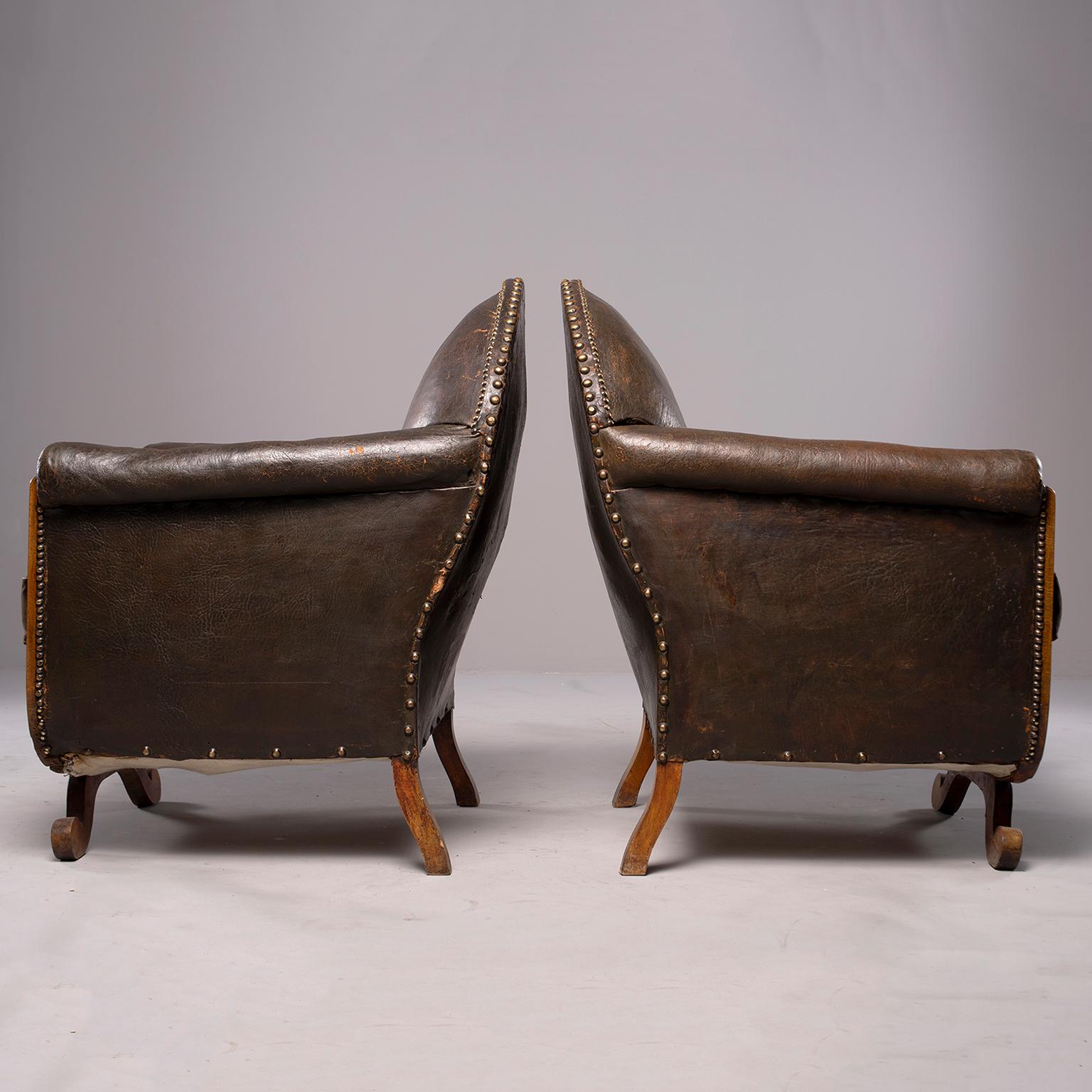 Pair of French leather club chairs with wood frames, circa 1930s. Camel back chairs with rolled arms, exposed wood frames, nailhead trim and curved, scrolled front leg. Original dark brown leather upholstery with dark brown velvet seat cushions.