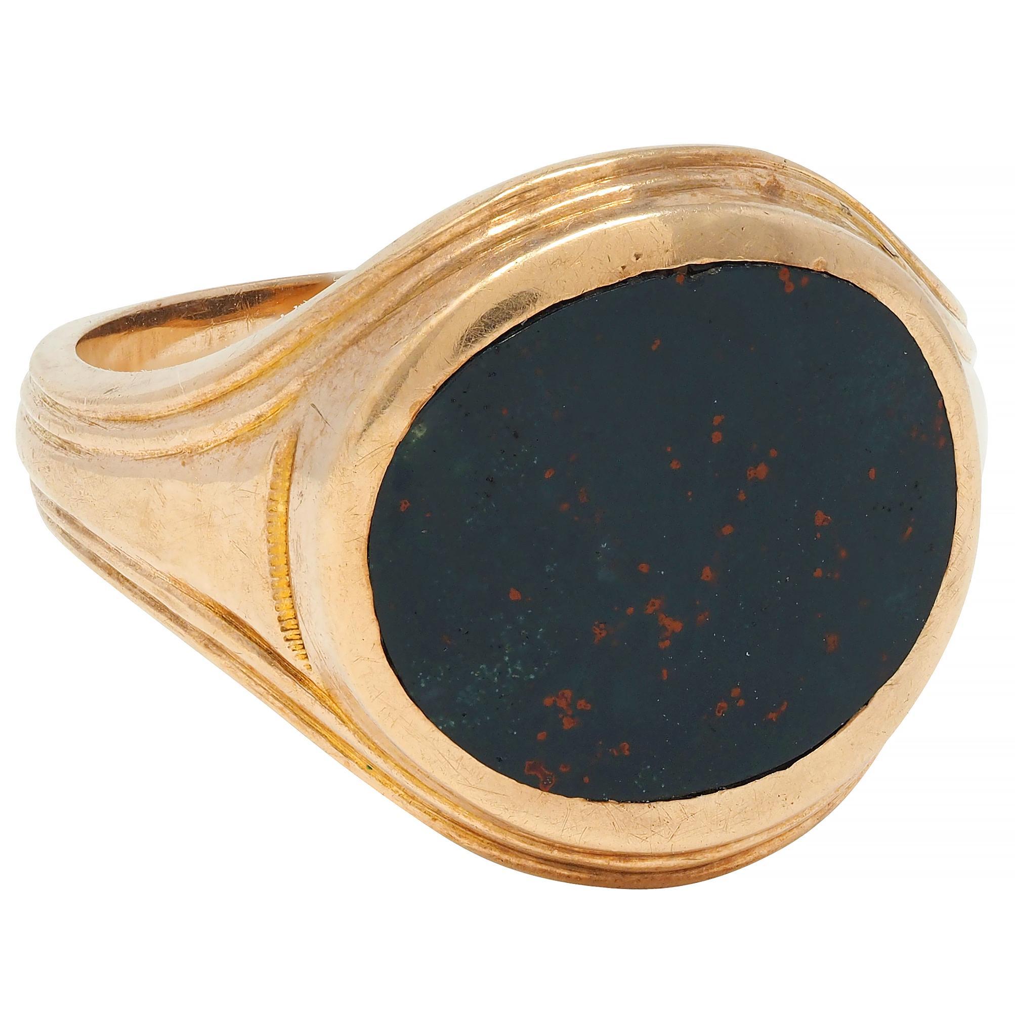 Centering an oval-shaped tablet of bloodstone measuring 12.0 x 14.0 mm 
Opaque dark bluish-green with orangy red flecking - bezel set east to west
Flanked by grooved shoulders with texture detail 
With ridged bombé shaped surround
Completed by