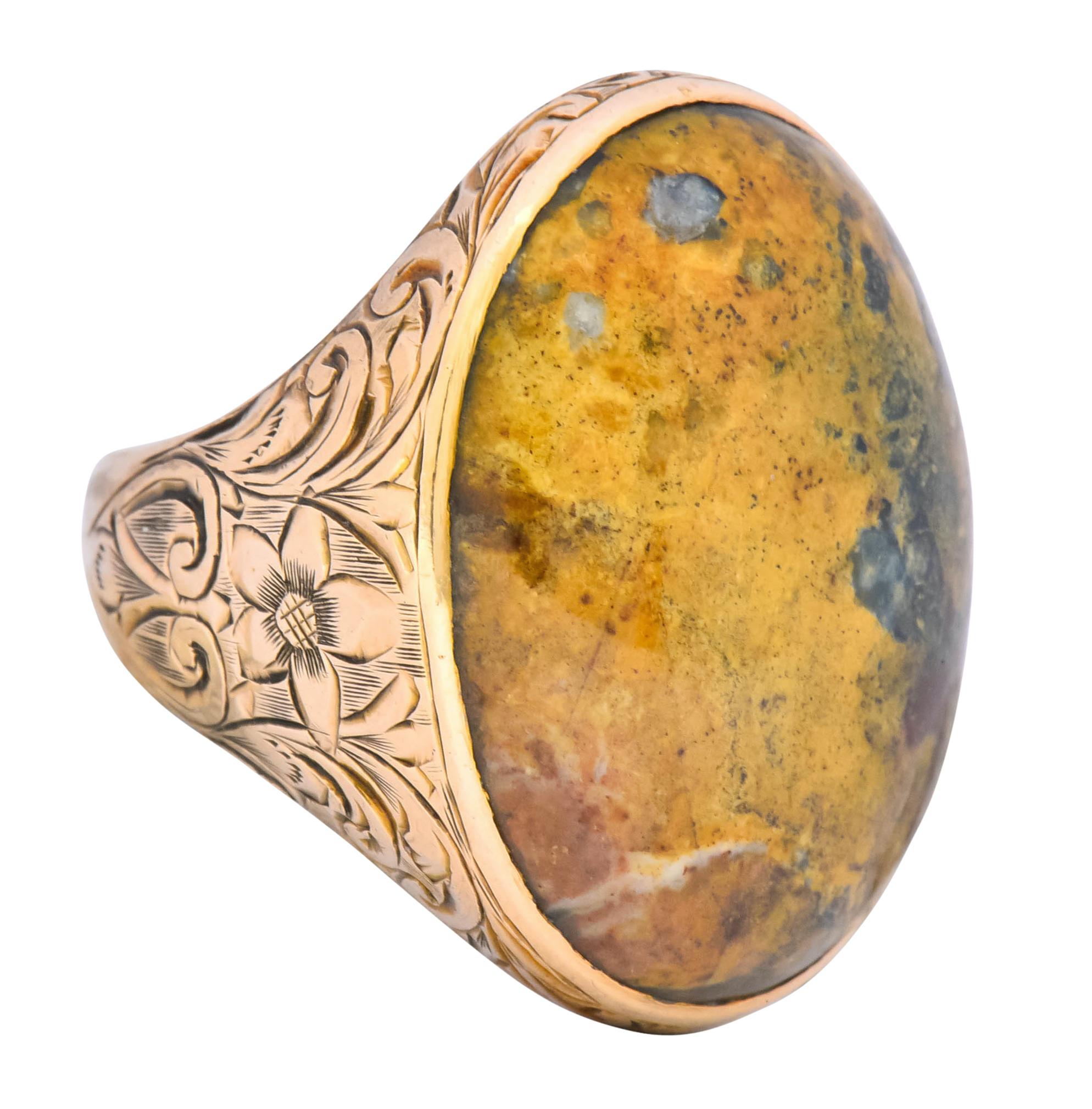 Centering an oval jasper cabochon measuring approximately 24.0 x 19.7 mm, opaque and marbled mustard yellow, rust red, and dark green

Bezel set in a decorative mounting deeply engraved with a scrolling foliate and floral motif

With maker's mark