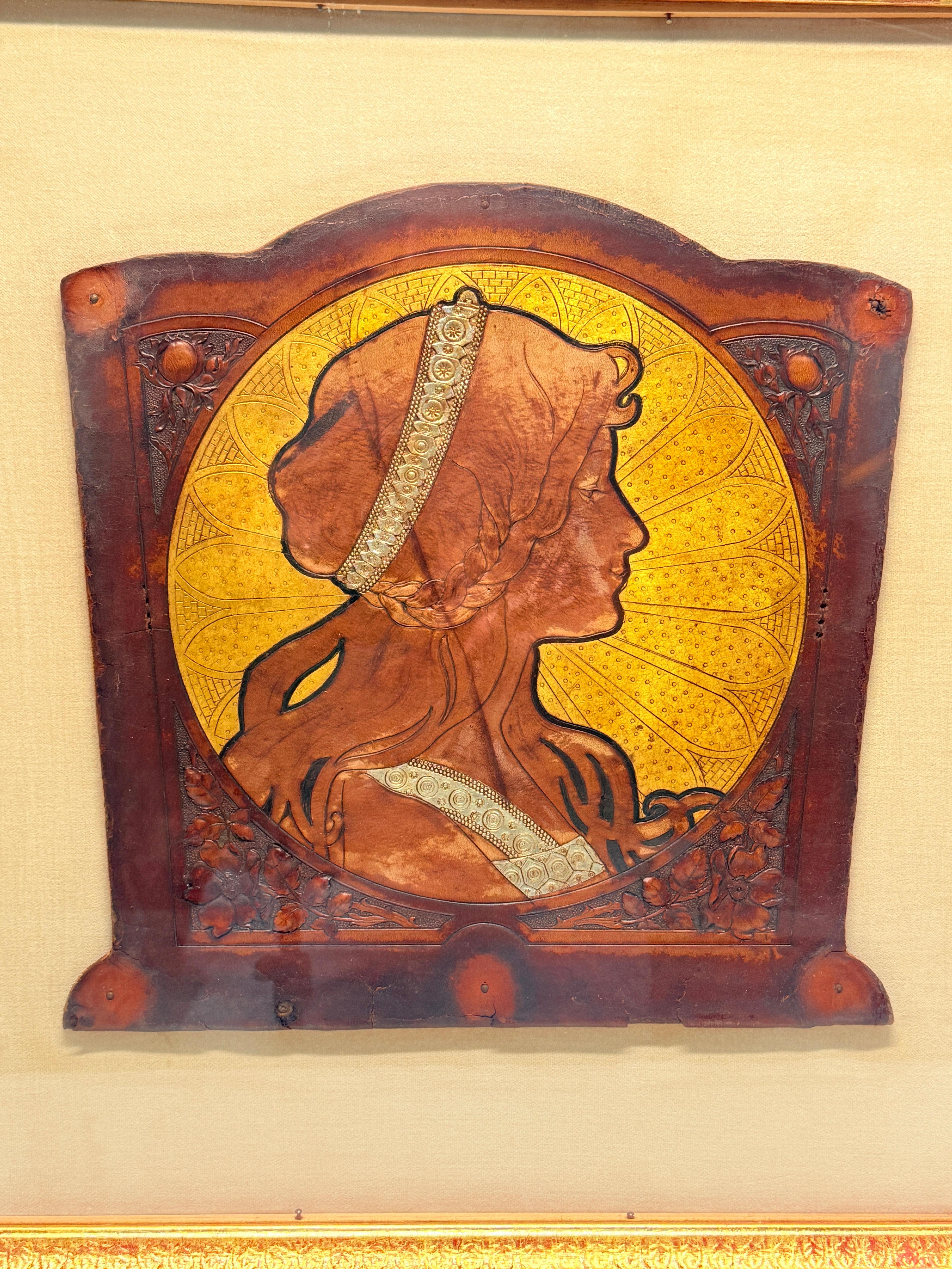 Un evocador panel de cuero repujado e inciso Art Nouveau atribuido a Alphonse Mucha, una de las figuras más influyentes de la Belle Époque. La composición presenta una romántica figura femenina de perfil, enmarcada en una radiante aureola dorada,
