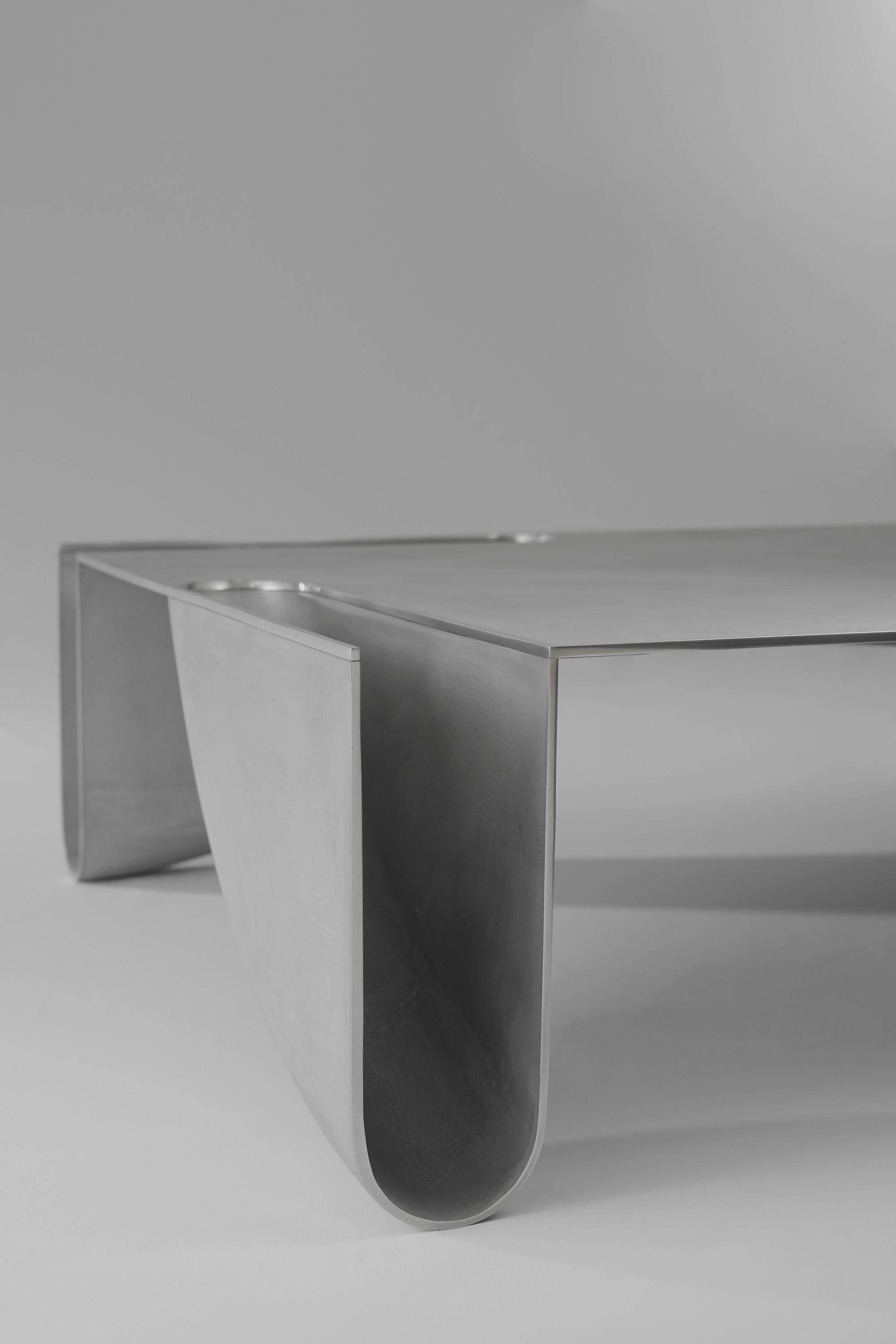 Une table basse contemporaine en métal moulé au sable avec des bords polis.
La table est composée de 4 éléments structurels de pieds coulés dans le sable, qui sont tous produits à l'aide du même moule et unis par le plateau de la table. Le métal est