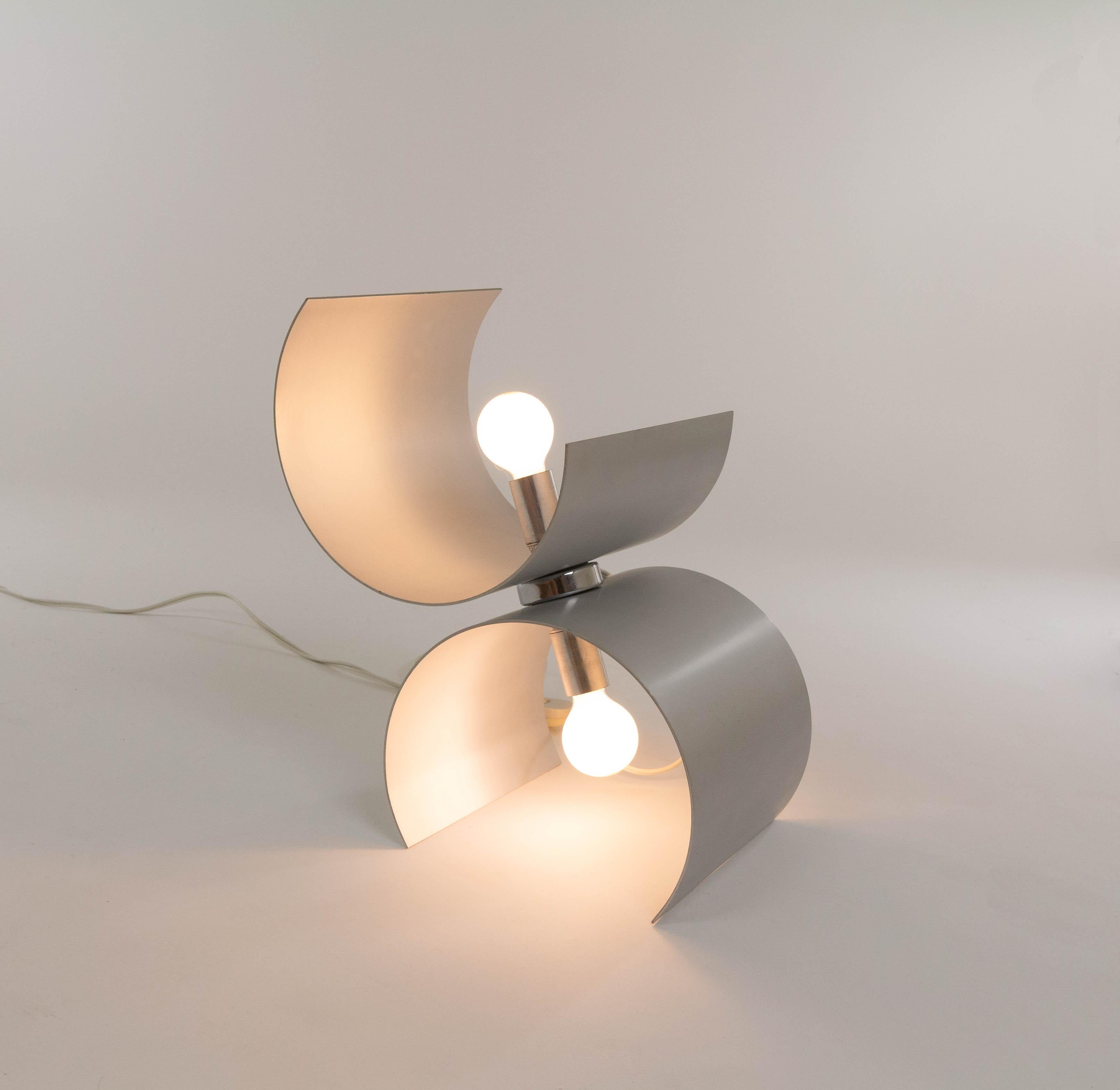 Table lamp produced by Nucleo Sormani in the 1970s.

The lamp consists of two curved aluminum parts, both containing a light bulb fitting (E14). The two parts are joined together, allowing the parts to rotate. The model of the curves offers a wide