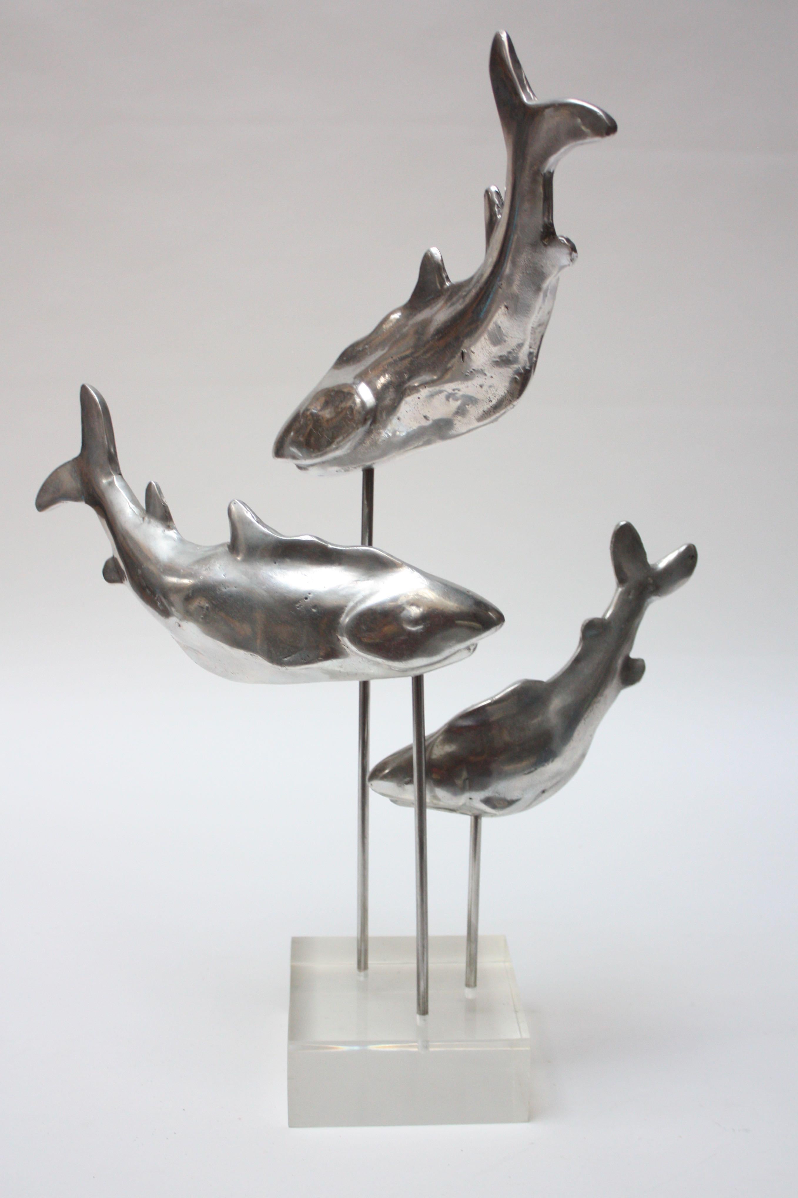 This unique sculpture is composed of three cast-aluminum sharks (representative of a family of sharks given the size disparity between the three) displayed in an ascending spiral formation on rods mounted onto a Lucite base. The sharks can removed