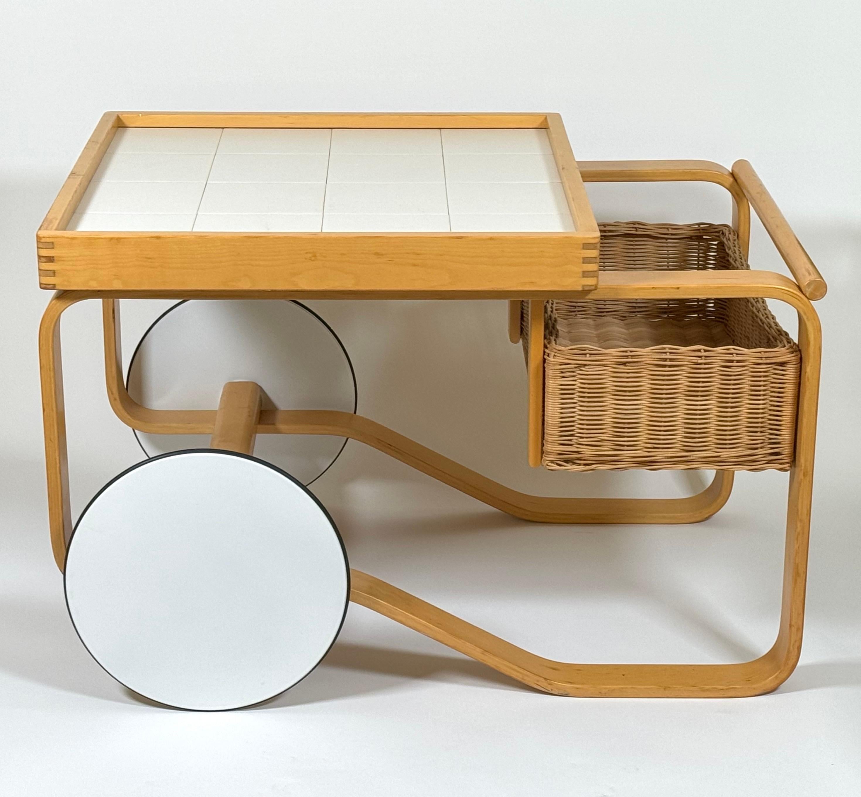 The Alvar and Aino Aalto 900 Trolley Tea / Bar Cart, originally designed in 1937, this is a 1980s production piece. Having a white ceramic tile top with a rattan basket on a bent wood birch frame with white lacquered and rubber edged wheels. The