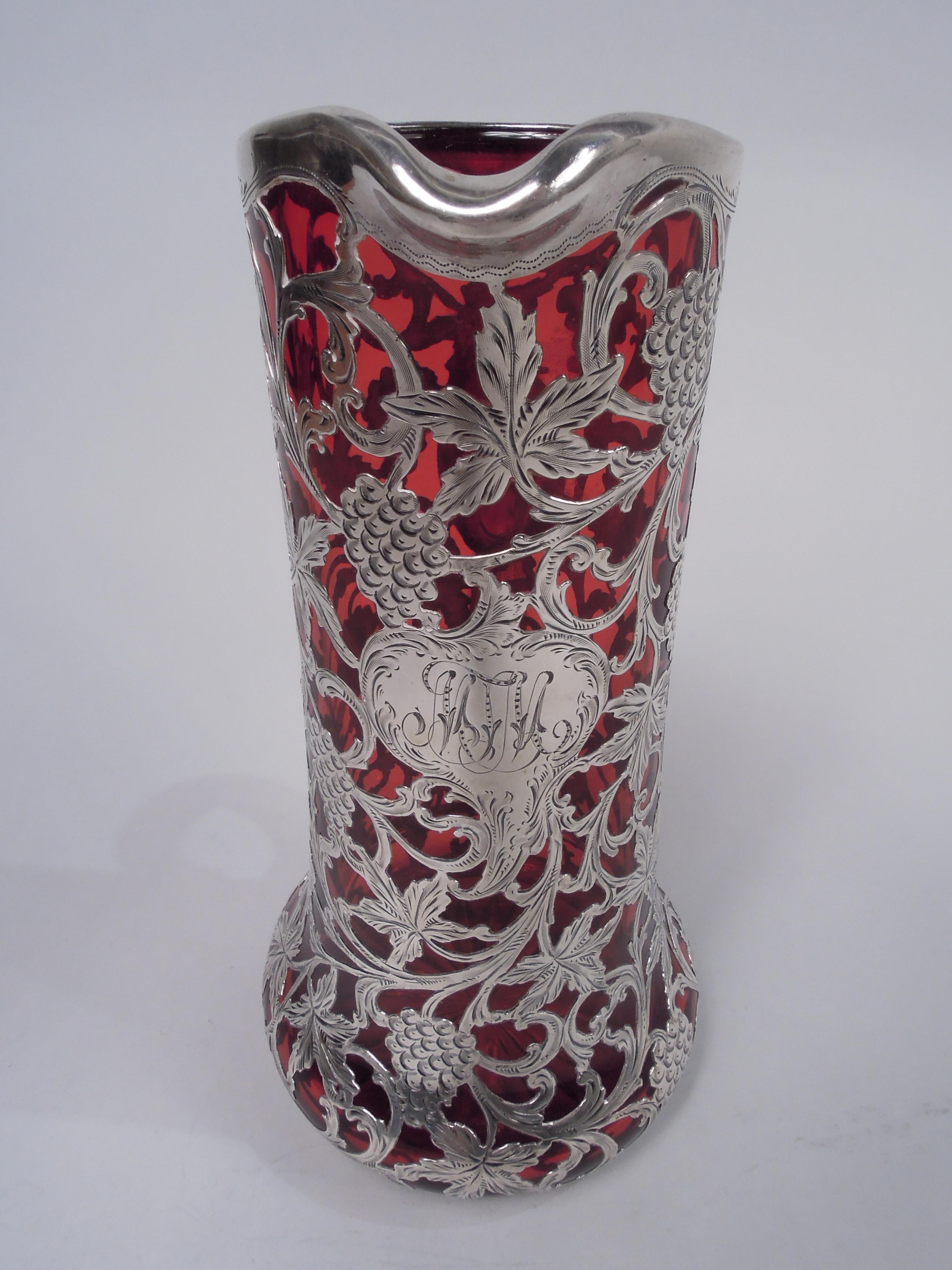 Art Nouveau glass claret jug with engraved silver overlay. Made by Alvin Corp. in Providence, ca 1900. Cylindrical with small lip spout and spread base; c-scroll handle in silver collar. On underside cut-to-clear star. Overlay in form of entwined