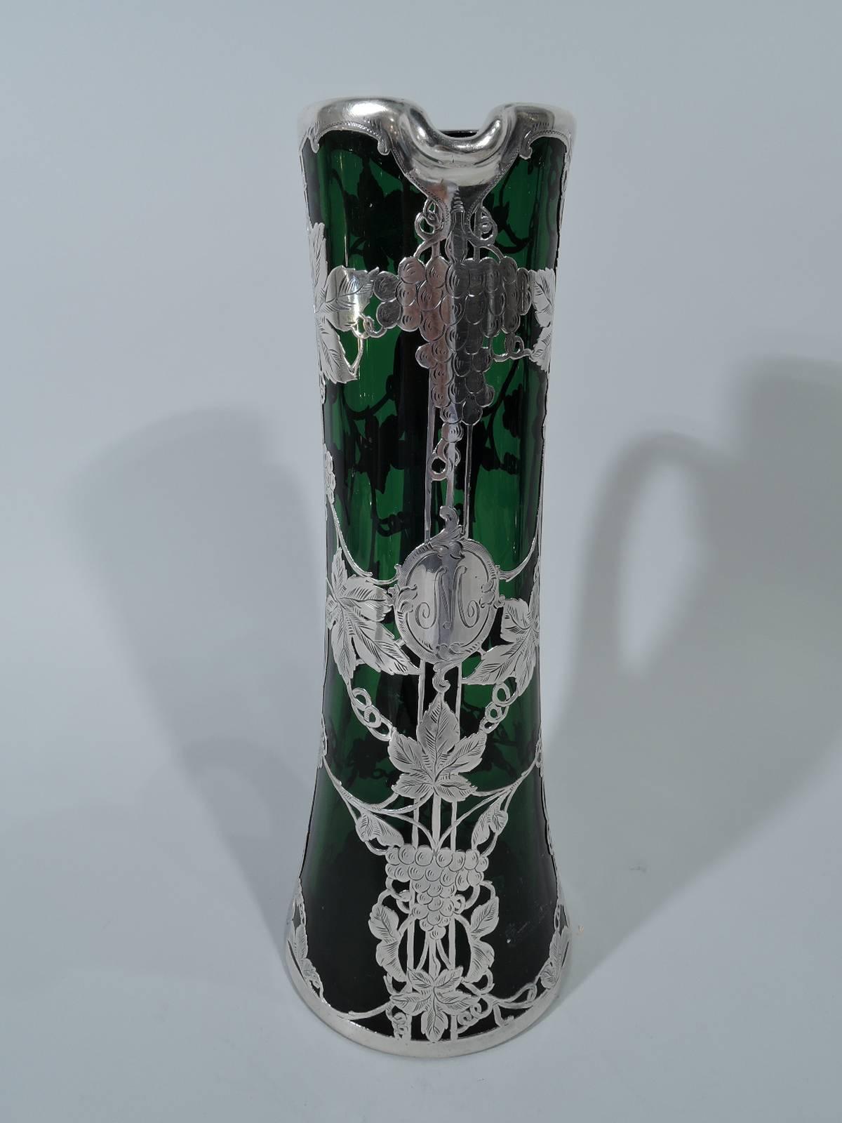 Art Nouveau emerald glass claret jug with silver overlay. Made by Alvin in Providence. Cylindrical with spread base and c-scroll handle. Overlay in form of vertical linear frame entwined with fruiting grapevines. Scrolled cartouche engraved with