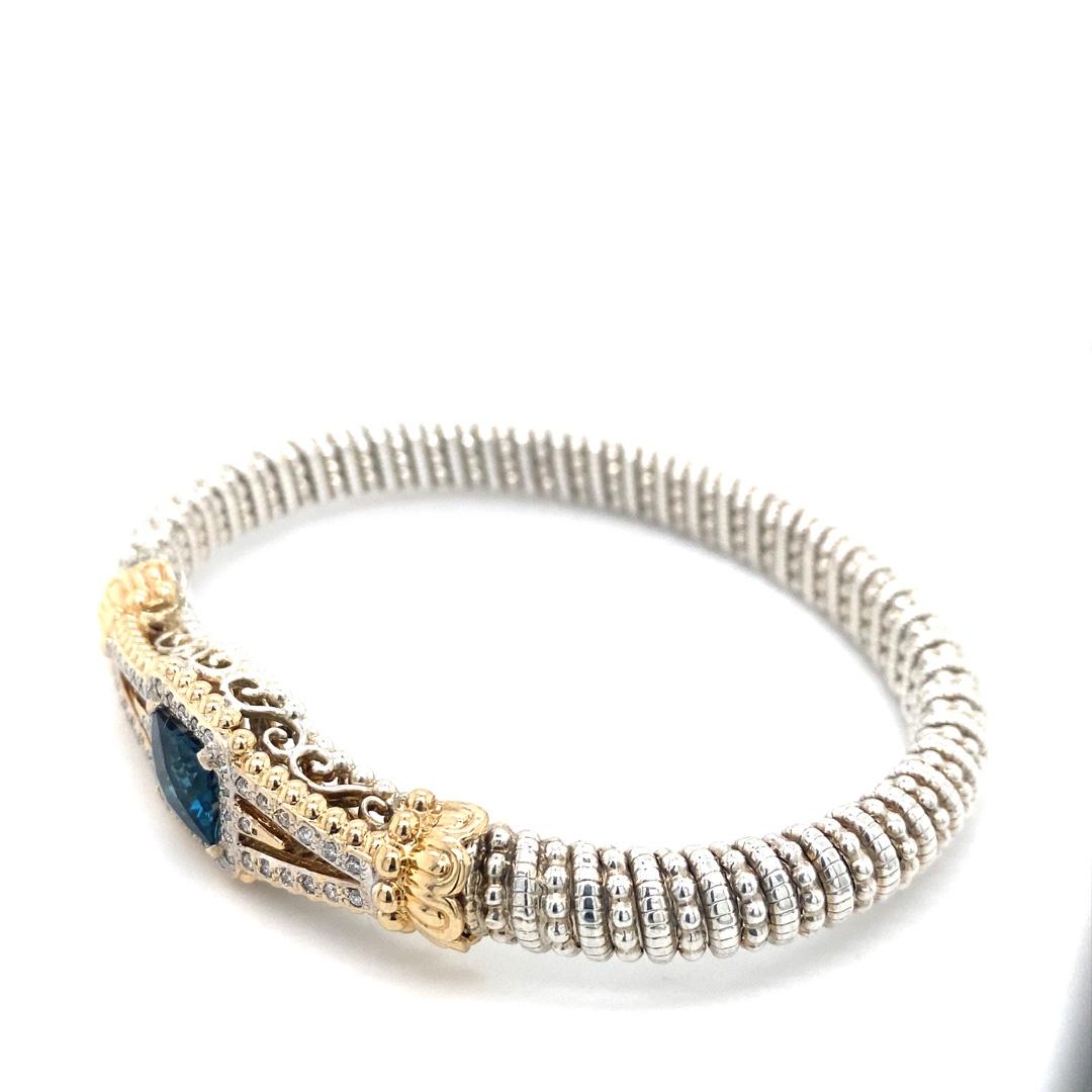 This handmade bracelet is designed by Alwand Vahan. There is 0.46cttw of diamonds set in 14 karat yellow gold. The bracelet is 8mm wide. The center stone is a London blue topaz. The bracelet features a beaded pattern in sterling silver, fitting a