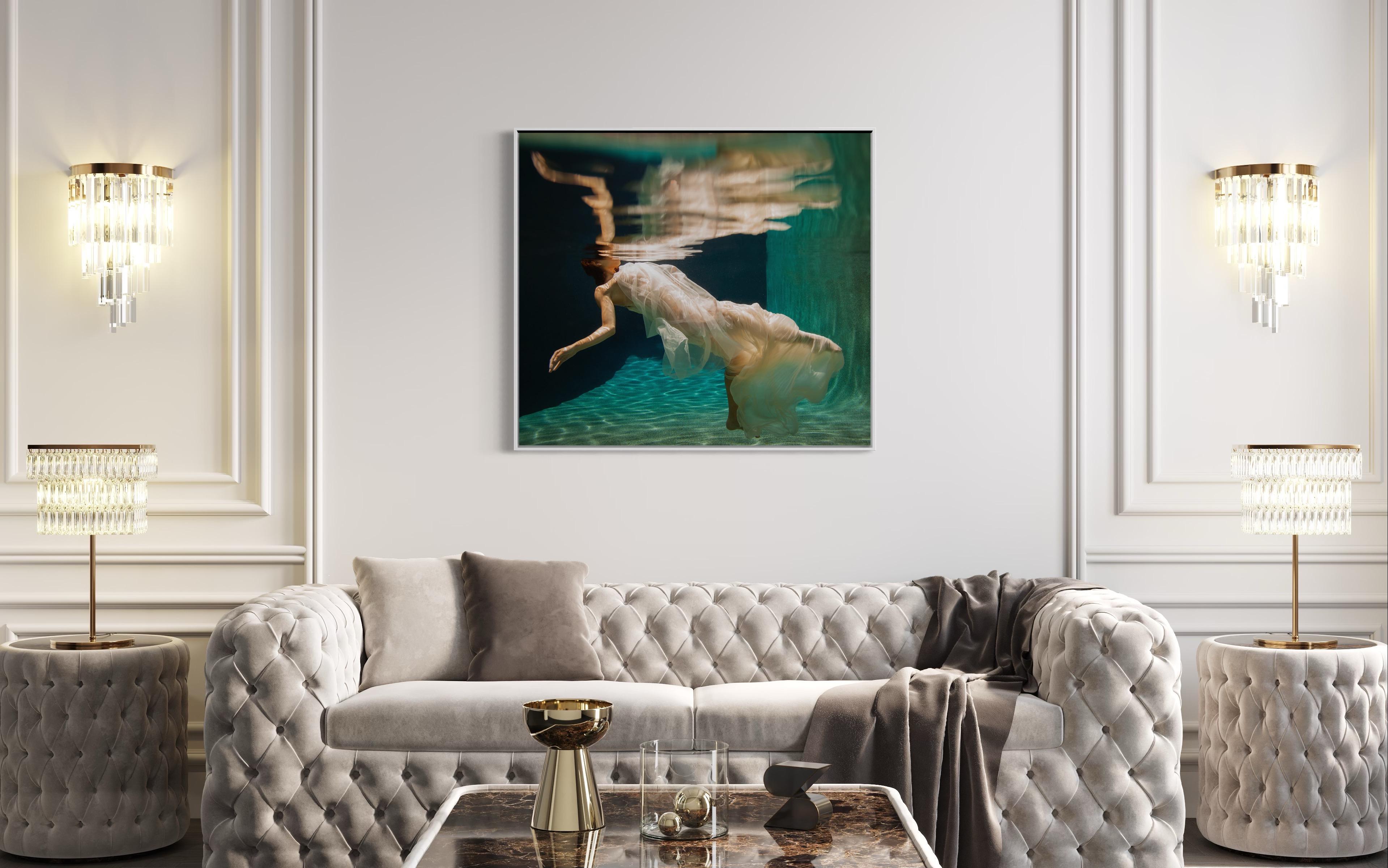 This contemporary figural limited edition fine art photograph by Alyssa Fortin captures a female figure wrapped in soft, light, creme-colored fabric, floating beneath the surface of water, with her face just above the surface and not visible to the