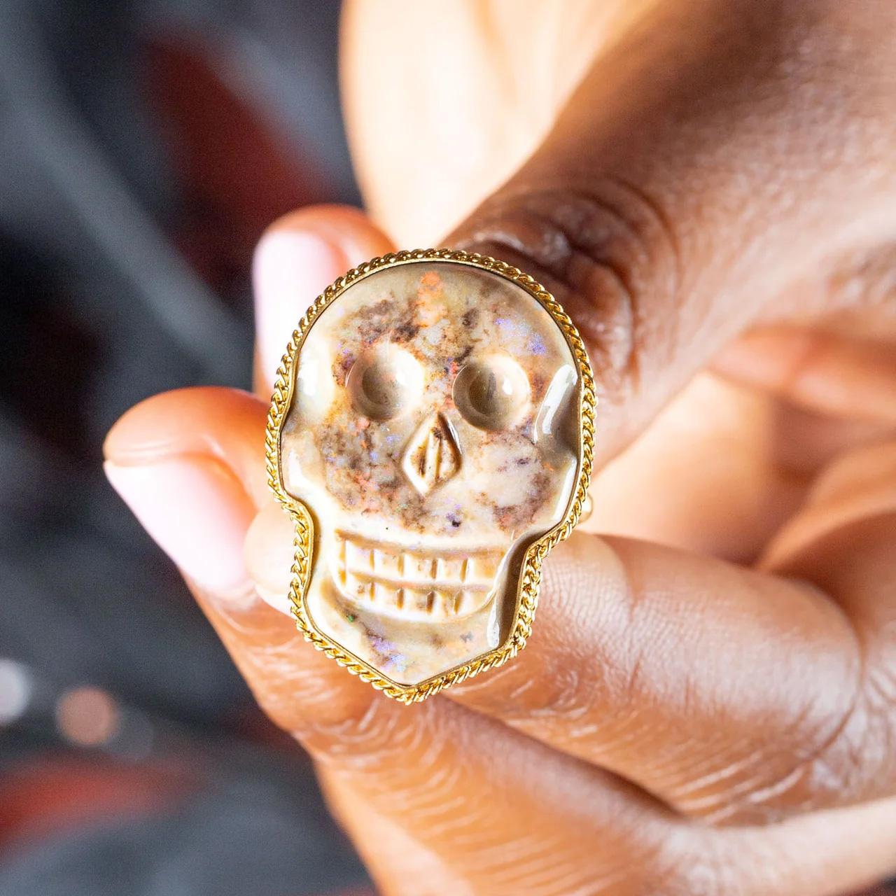 Glints of red, green, and blue dance across the surface of this carved Andamooka Opal skull. Blemishes of brown and black add to the hauntingly realistic appearance of this spooky skull ring. Set in an 18k gold bezel with braided detail.

Ready to