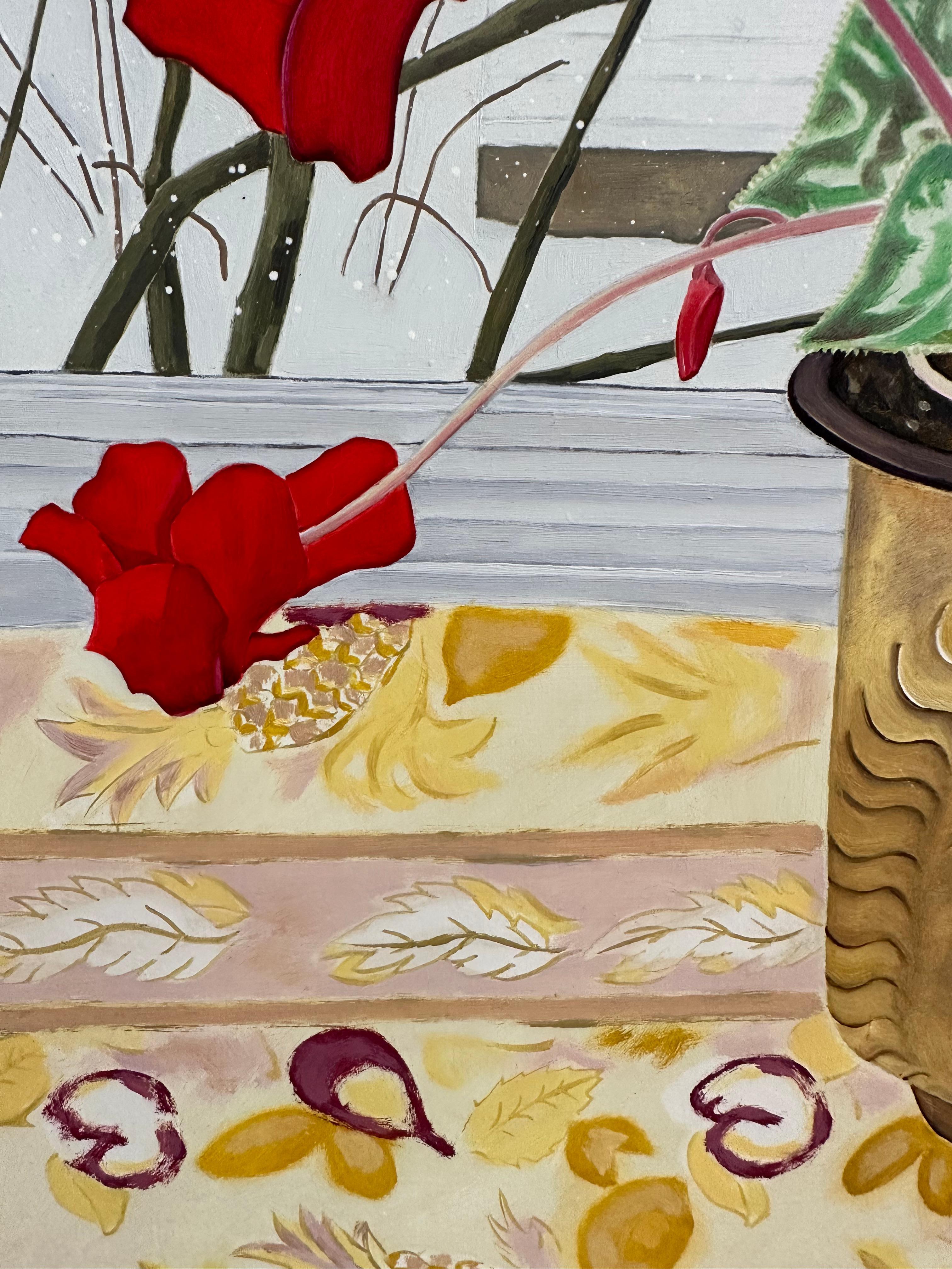 Crimson red flowers with dark green leaves in a golden planter with a face on the side sit on a patterned yellow tablecloth in front of a window. Through the window, a winter landscape with freshly falling snow and a white house can be seen outside.