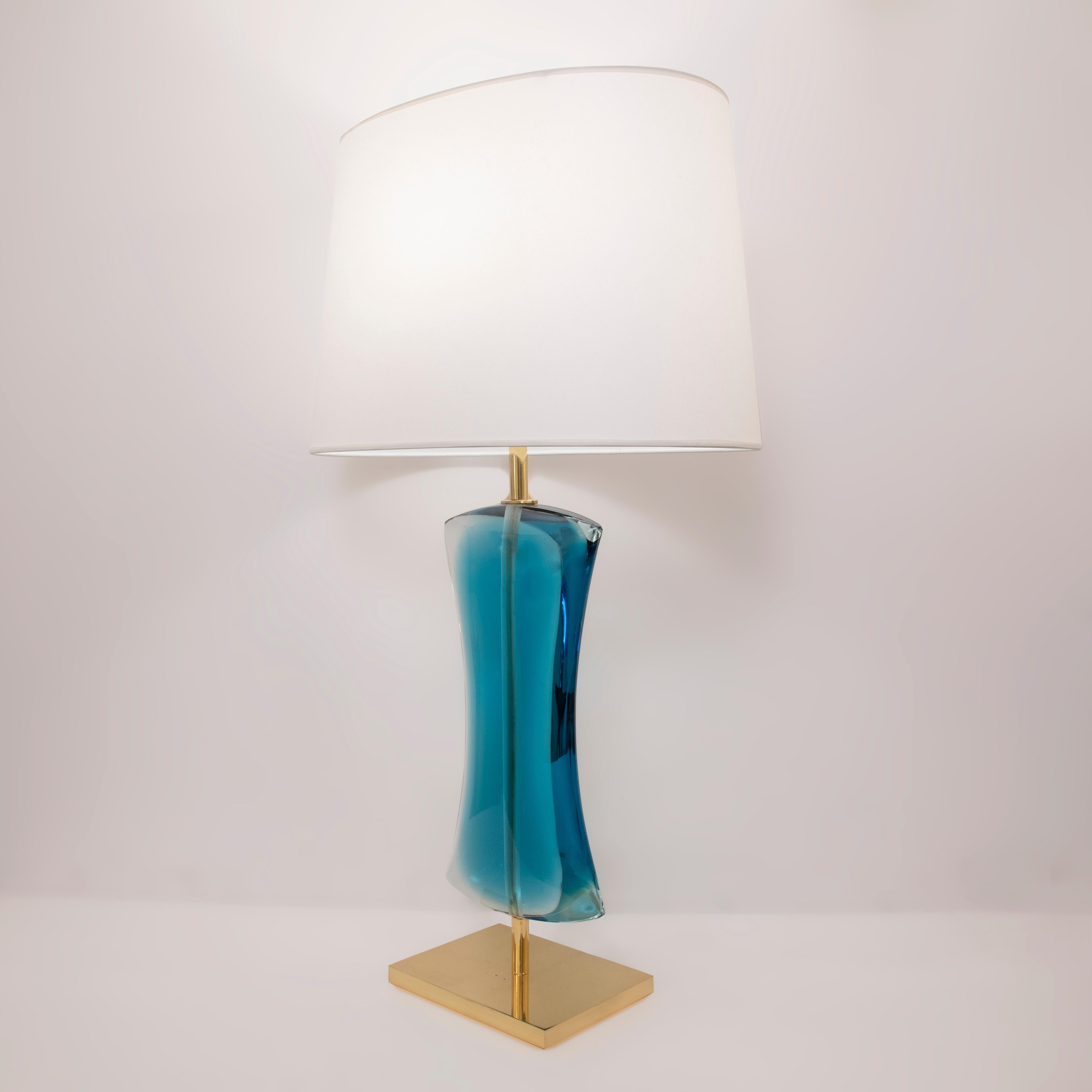 The Amante table lamp is minimalist representation of the human body with its sensual and curvaceous lines. Fabricated by several glass sheets bound together and then carved by hand. Shown in polished brass with blue glass. Shade not