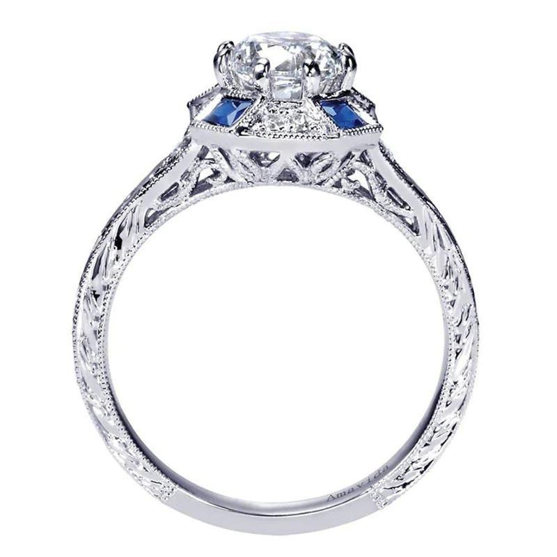 Vintage inspired diamond engagement ring in platinum with blue sapphire accents. Ring features delicate diamond etching on the sides, and a cushion shaped diamond halo with sapphire accents in the corners. Mounting contains 0.09 ctw of premium white