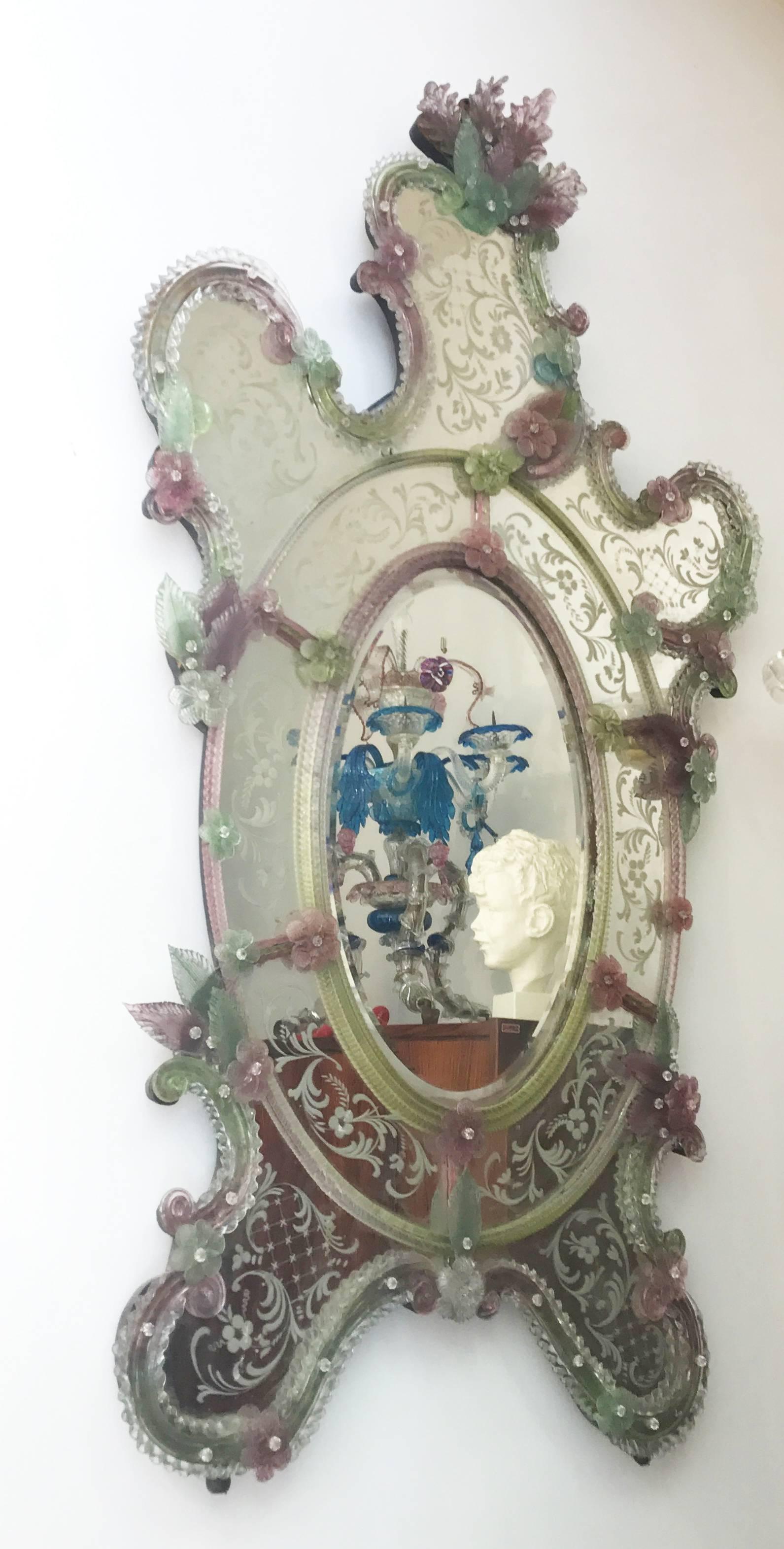 This beautiful Venetian mirror features etched floral motifs adorning the mirrored frame. Along the edges of the frame are glass rope accents and numerous glass pink and green flowers.
Very good vintage condition.
  