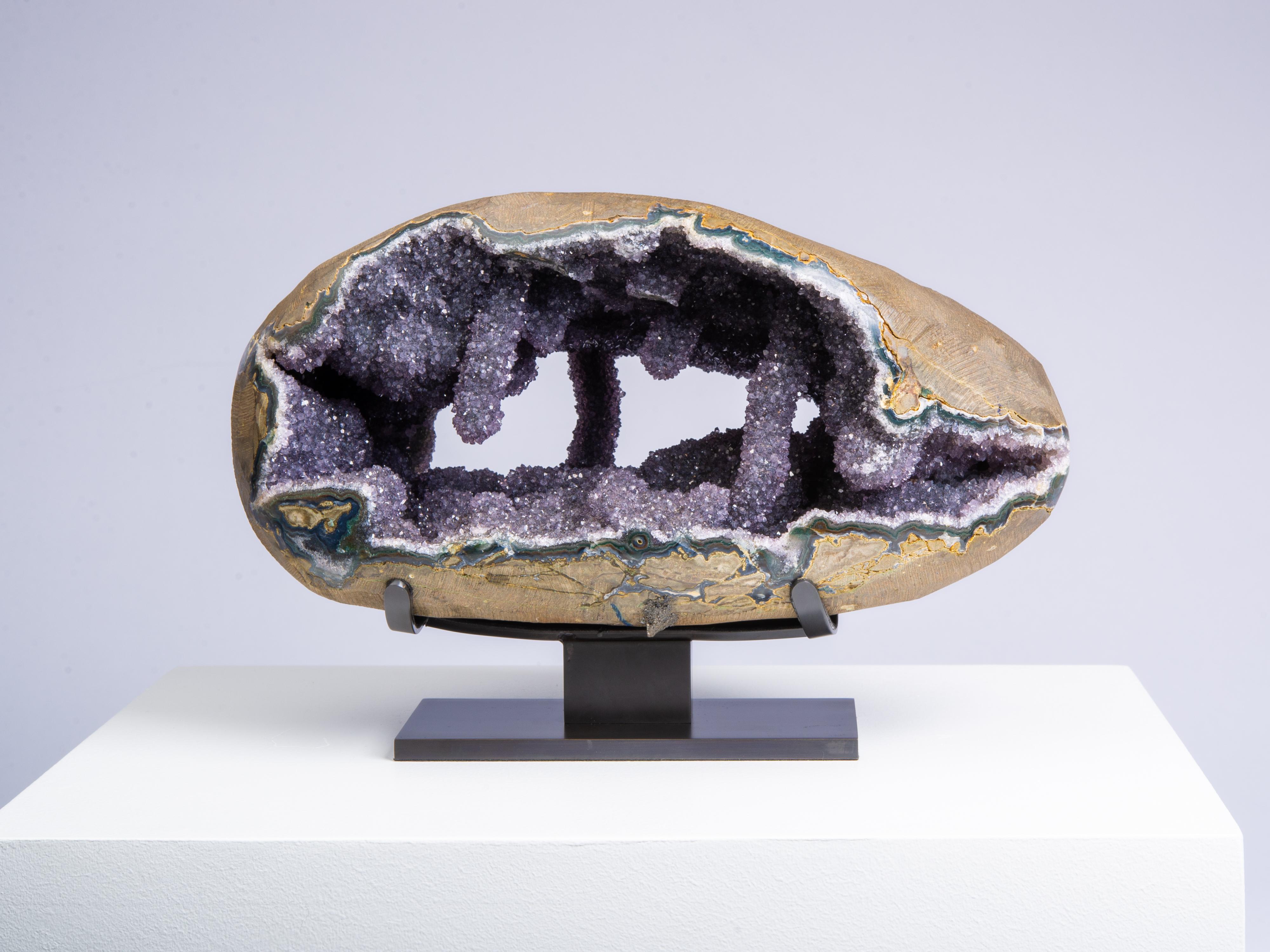 This extraordinary piece is a stunning geode displayed stylishly on a decorative a wooden stand. The interior of the geode comprising a dark grey amethyst Druze with multiple stalactite and stalagmite formations. The outer layer of the piece is in